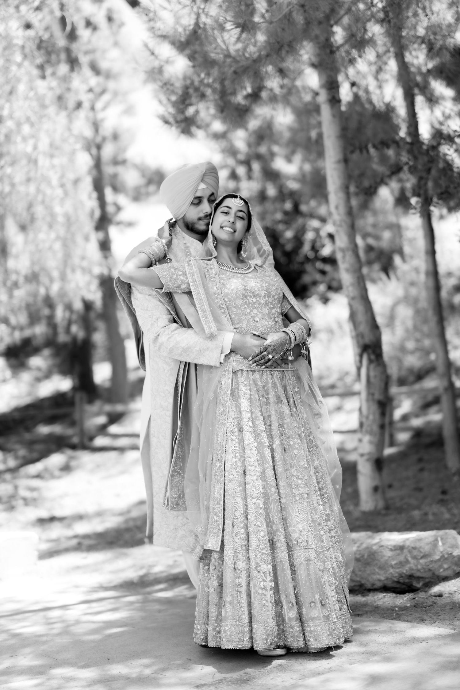 Black and white photo of a Sikh bride and groom in traditional attire embracing outdoors among trees.