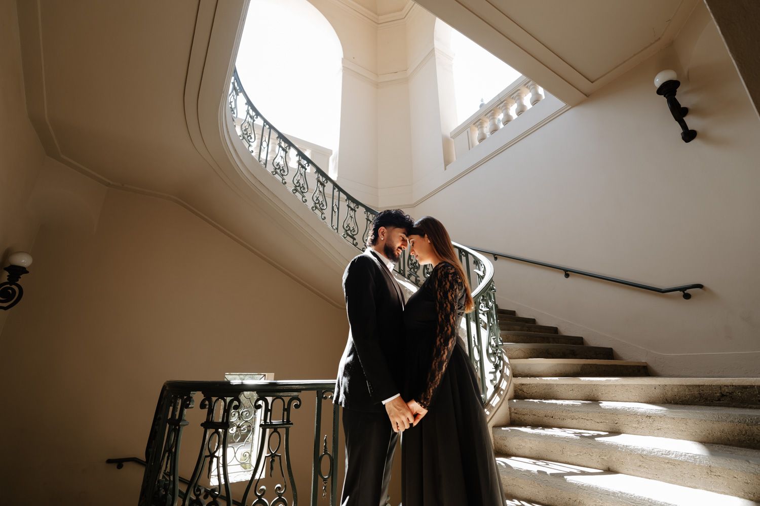 Couple in black attire share a romantic moment on an ornate grand staircase in an elegant venue.