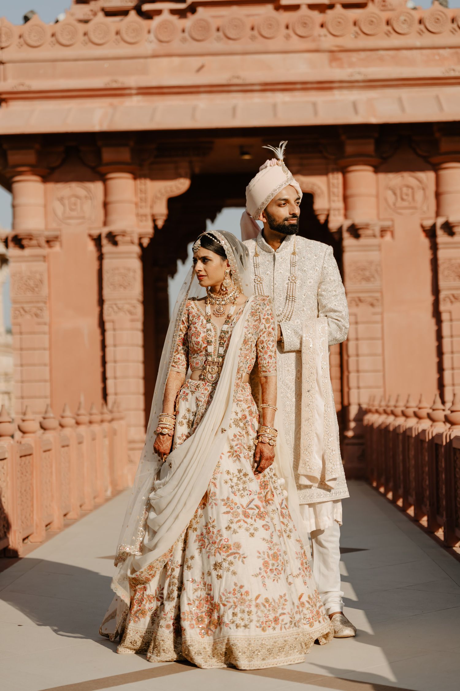 Couple in traditional Indian wedding attire standing in pink sandstone temple corridor.