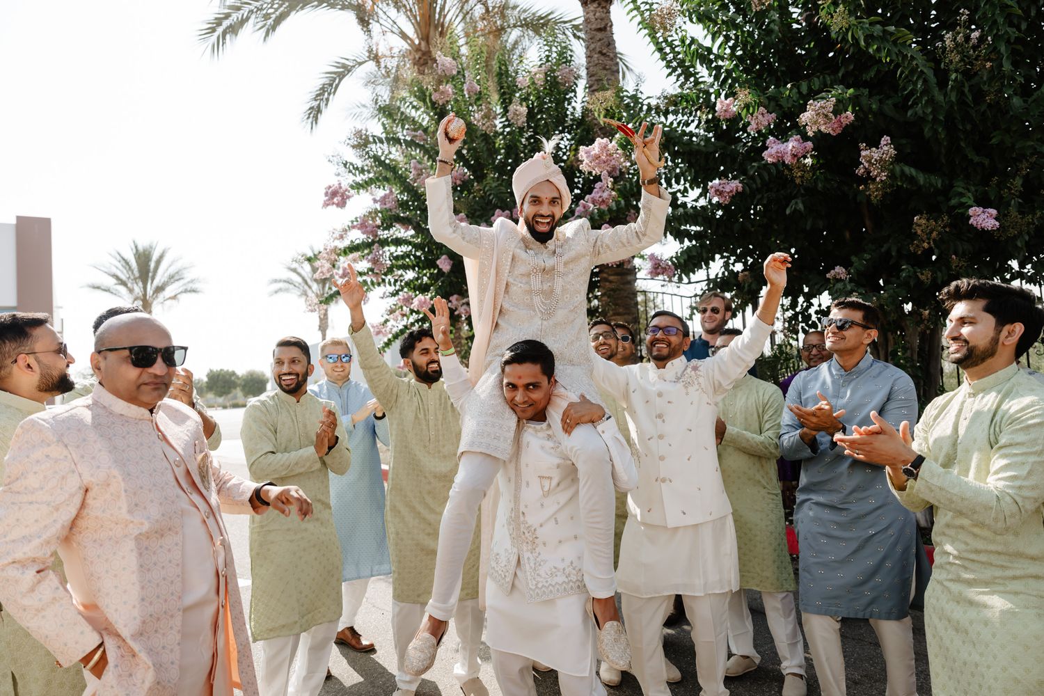 Wedding celebration with guests in light colored attire dancing and celebrating outdoors under palm trees.