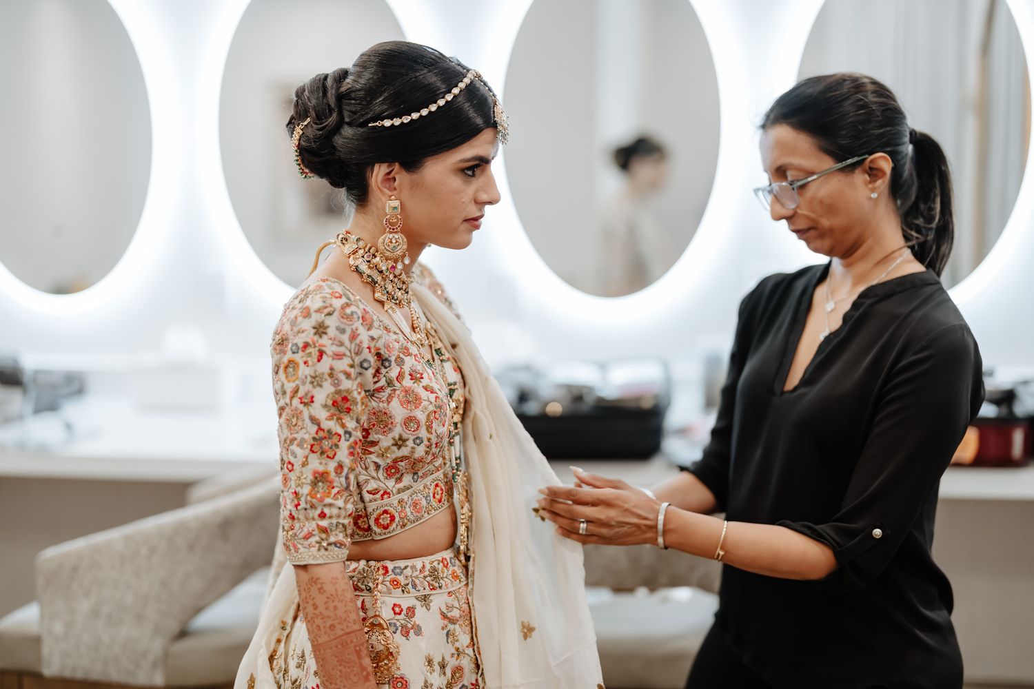 Bride and stylist prepare for wedding in elegant white room with ornate decor and natural lighting.