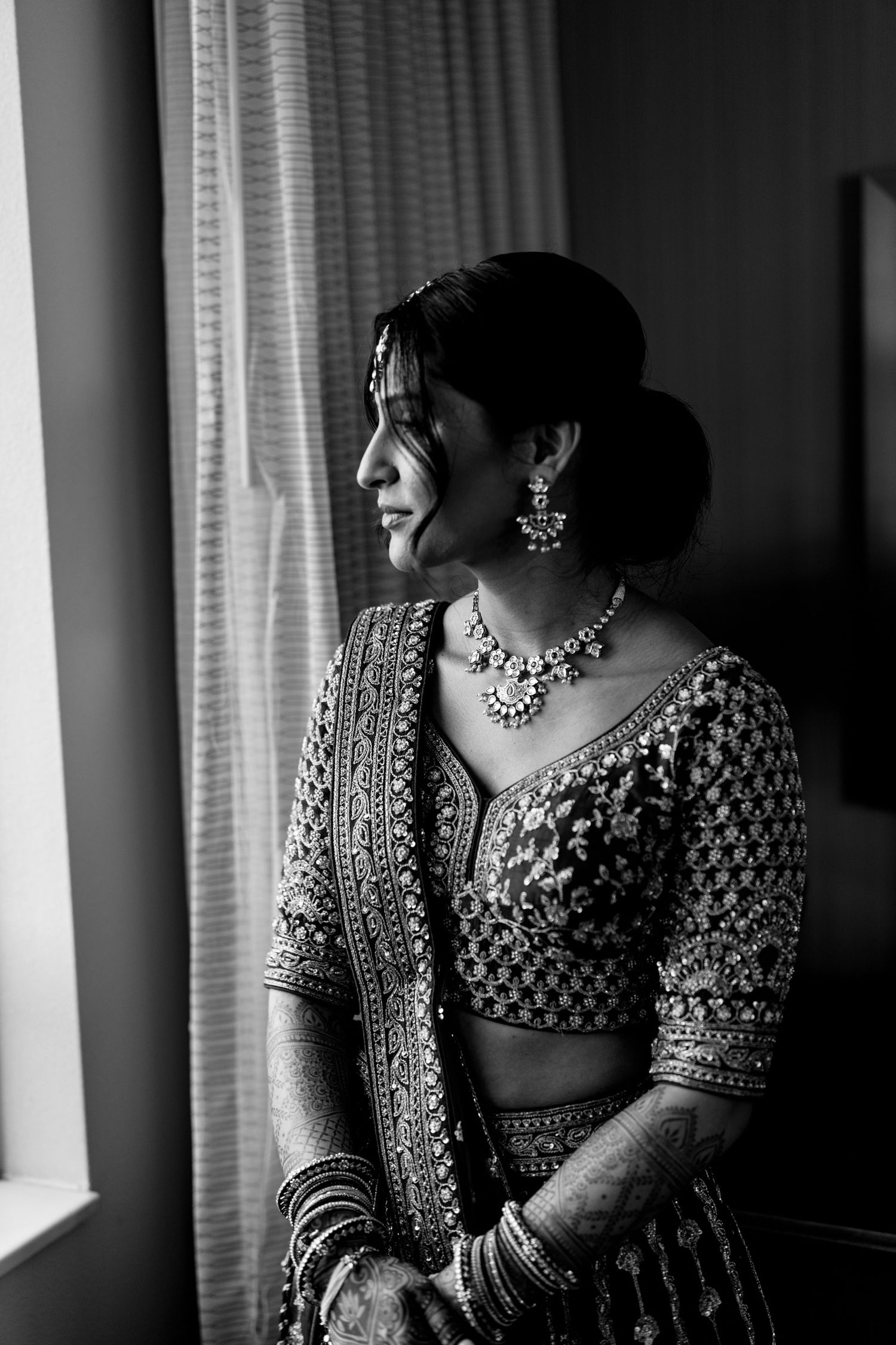A black and white portrait of someone in traditional Indian bridal attire with ornate jewelry looking towards a window.