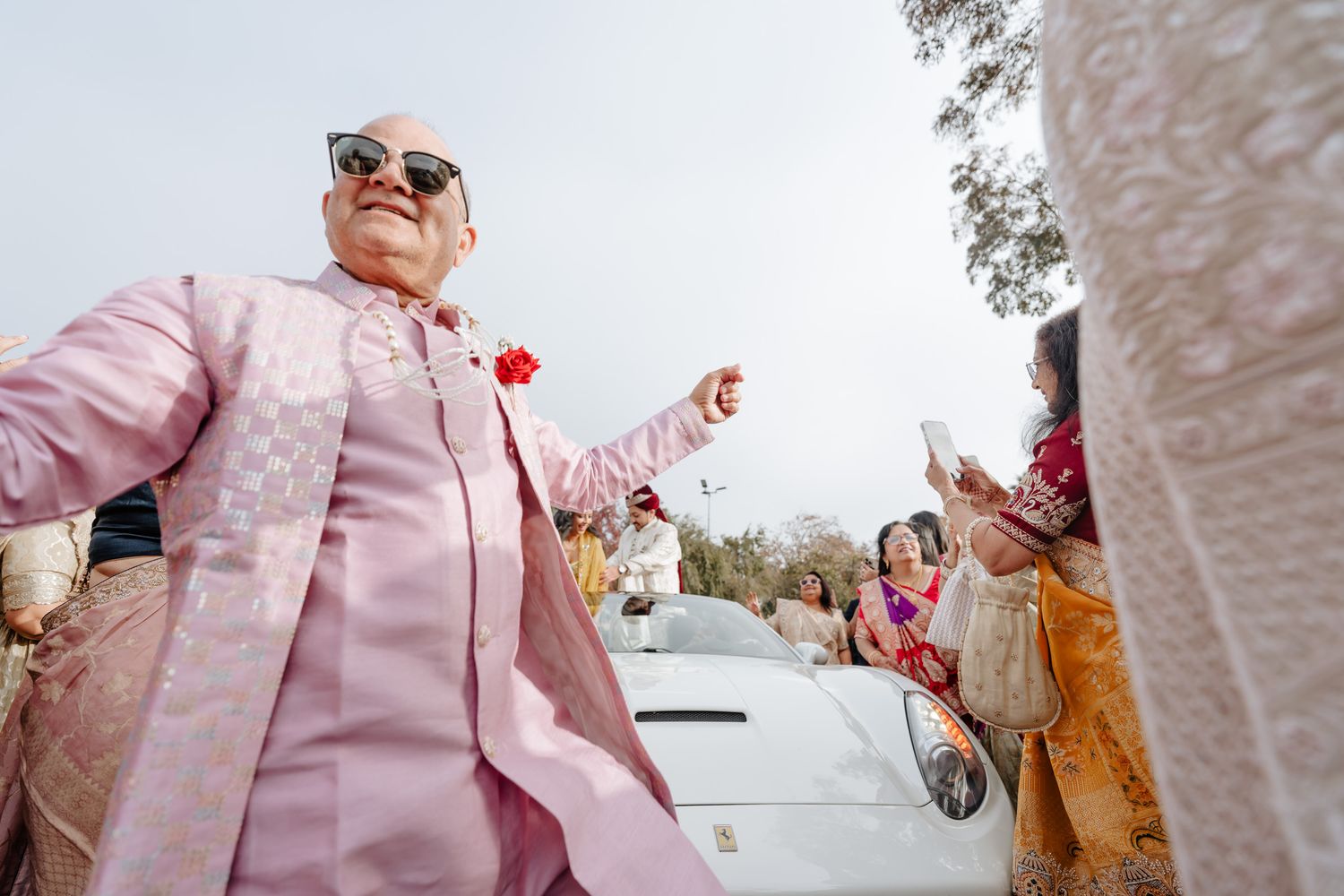 A person in a pink jacket dances joyfully outdoors with others in the background wearing orange clothing.