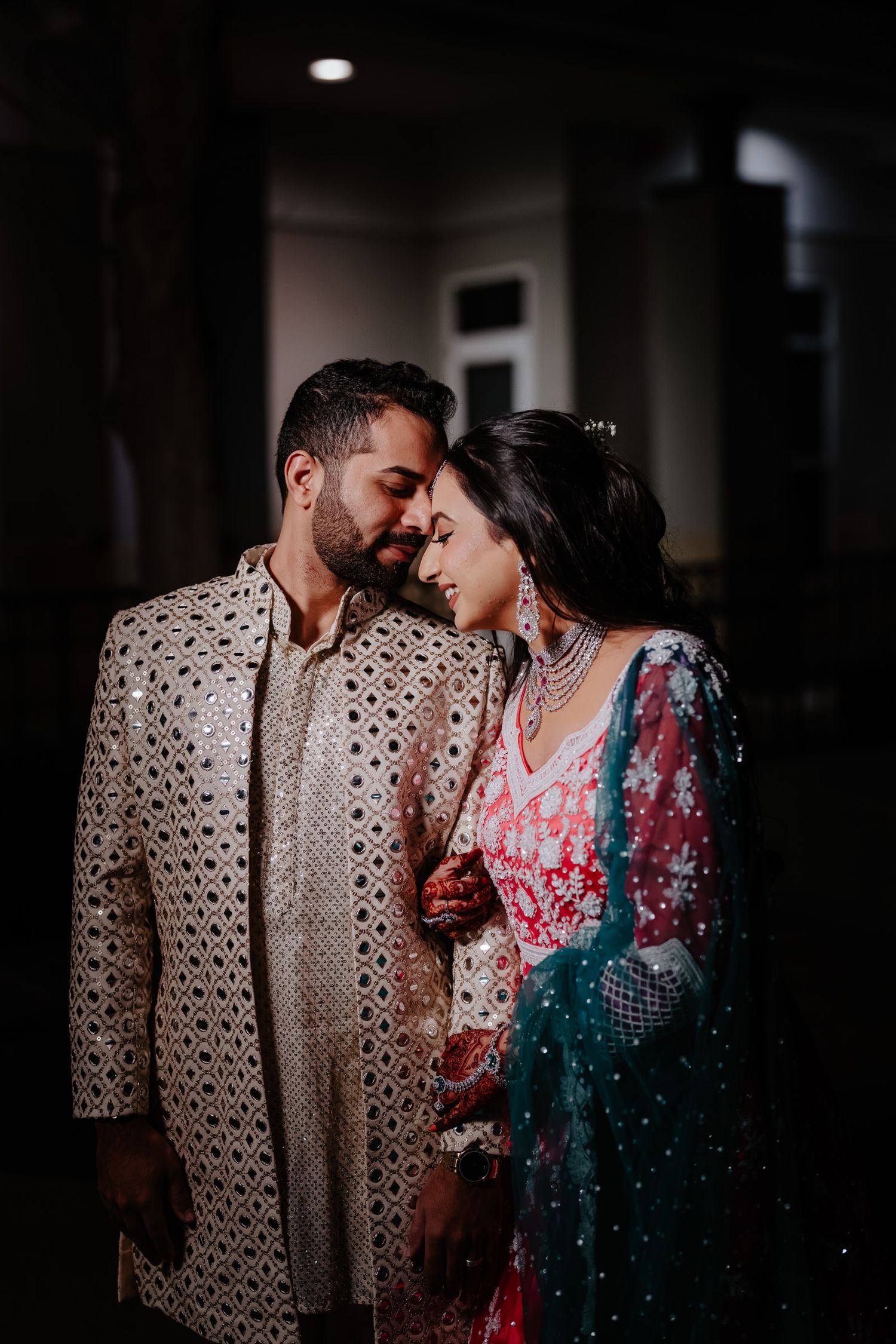 A couple in traditional South Asian wedding attire share an intimate moment in a dimly lit hallway.