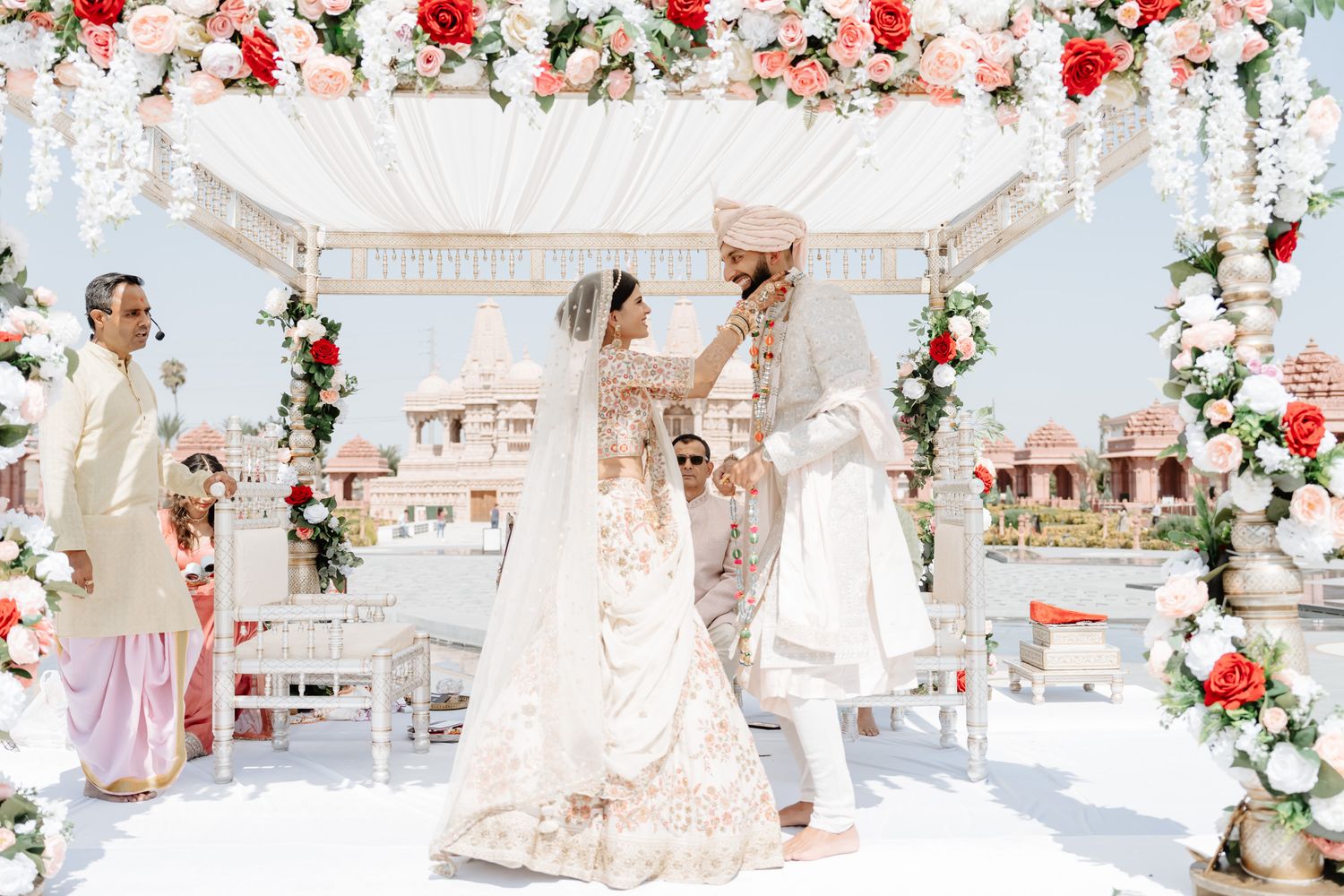 A romantic wedding ceremony setup with white drapes, floral arrangements, and red roses in an elegant venue.