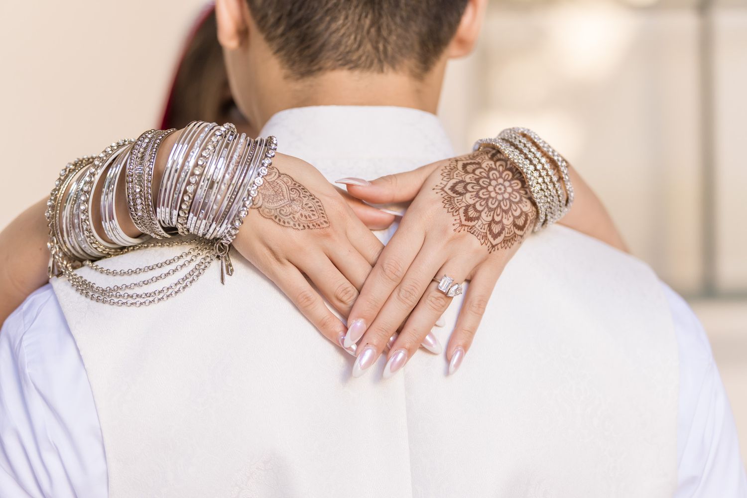 Hands adorned with intricate henna designs and silver bangles embrace across a white-clothed shoulder.