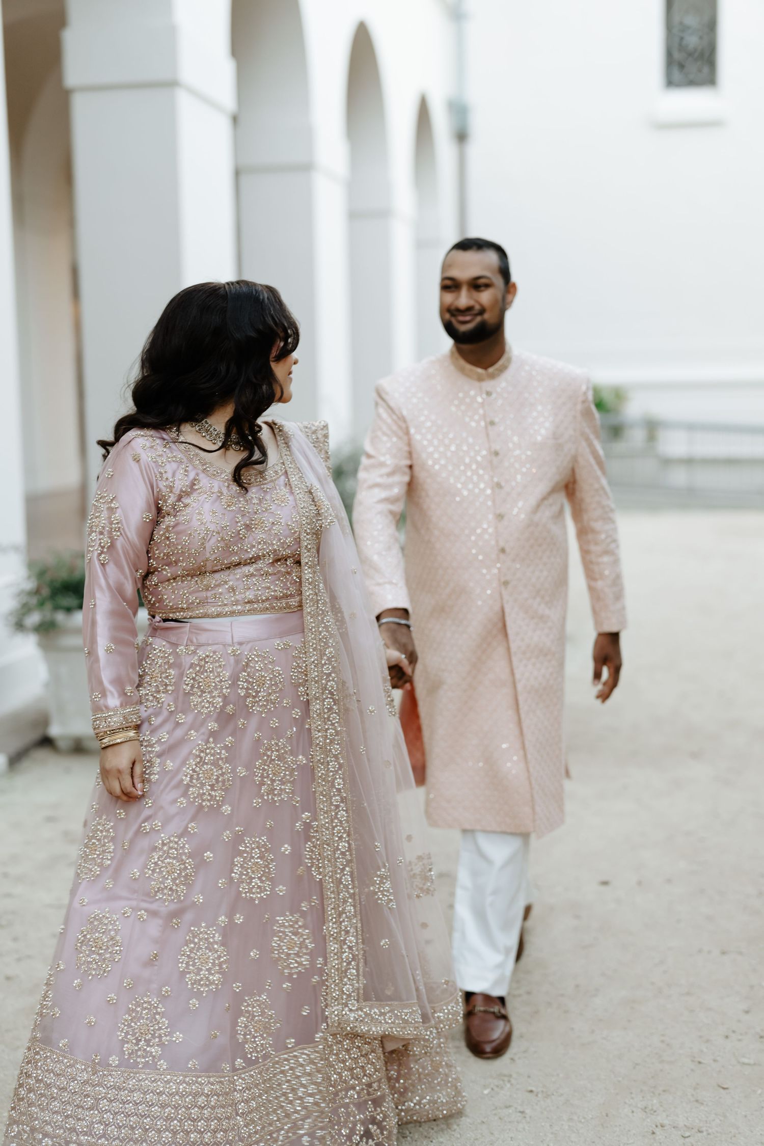 A couple in matching pink traditional Indian wedding attire walking along a colonnade.