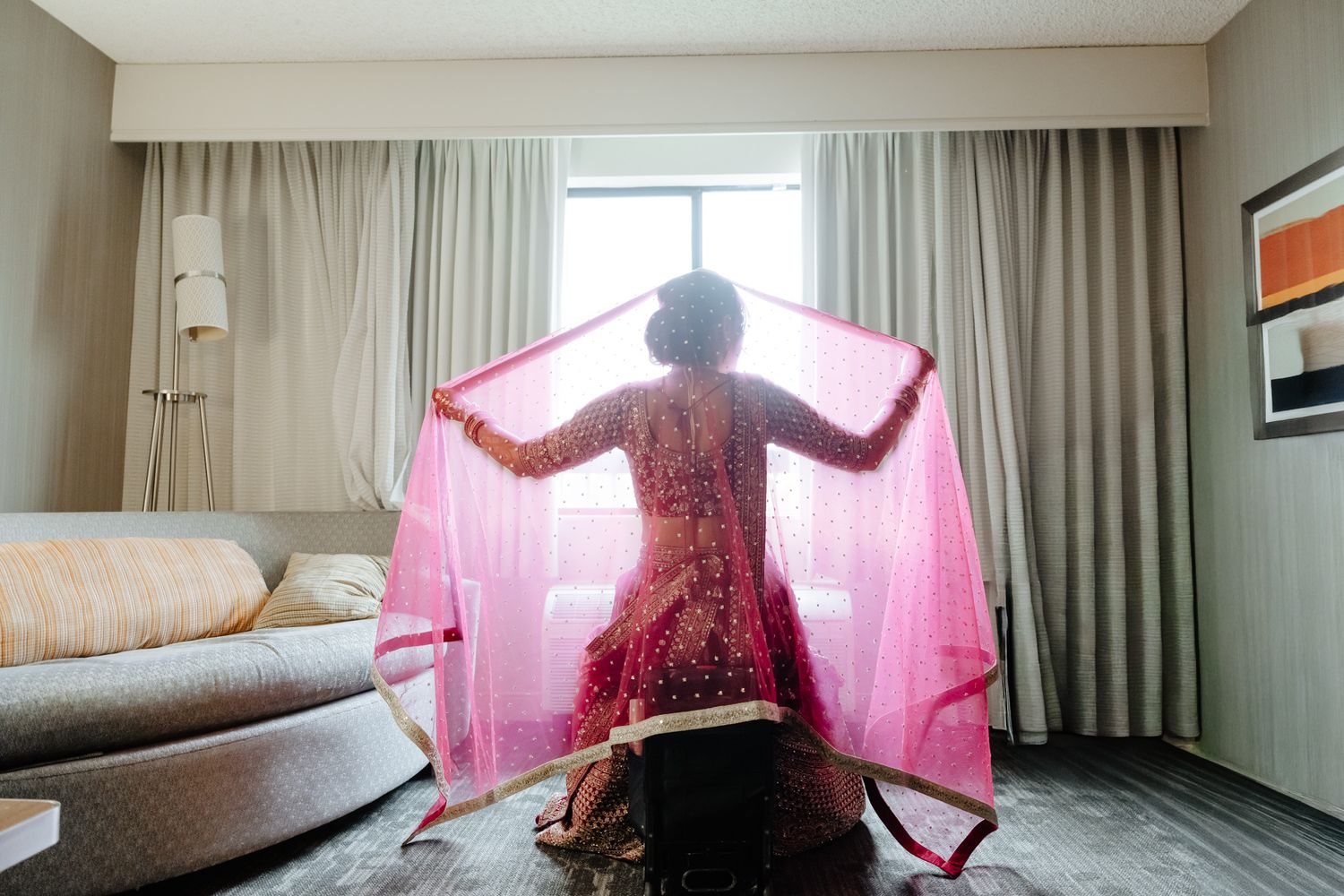 A silhouette in a flowing pink dress stands backlit by window light in a hotel room.