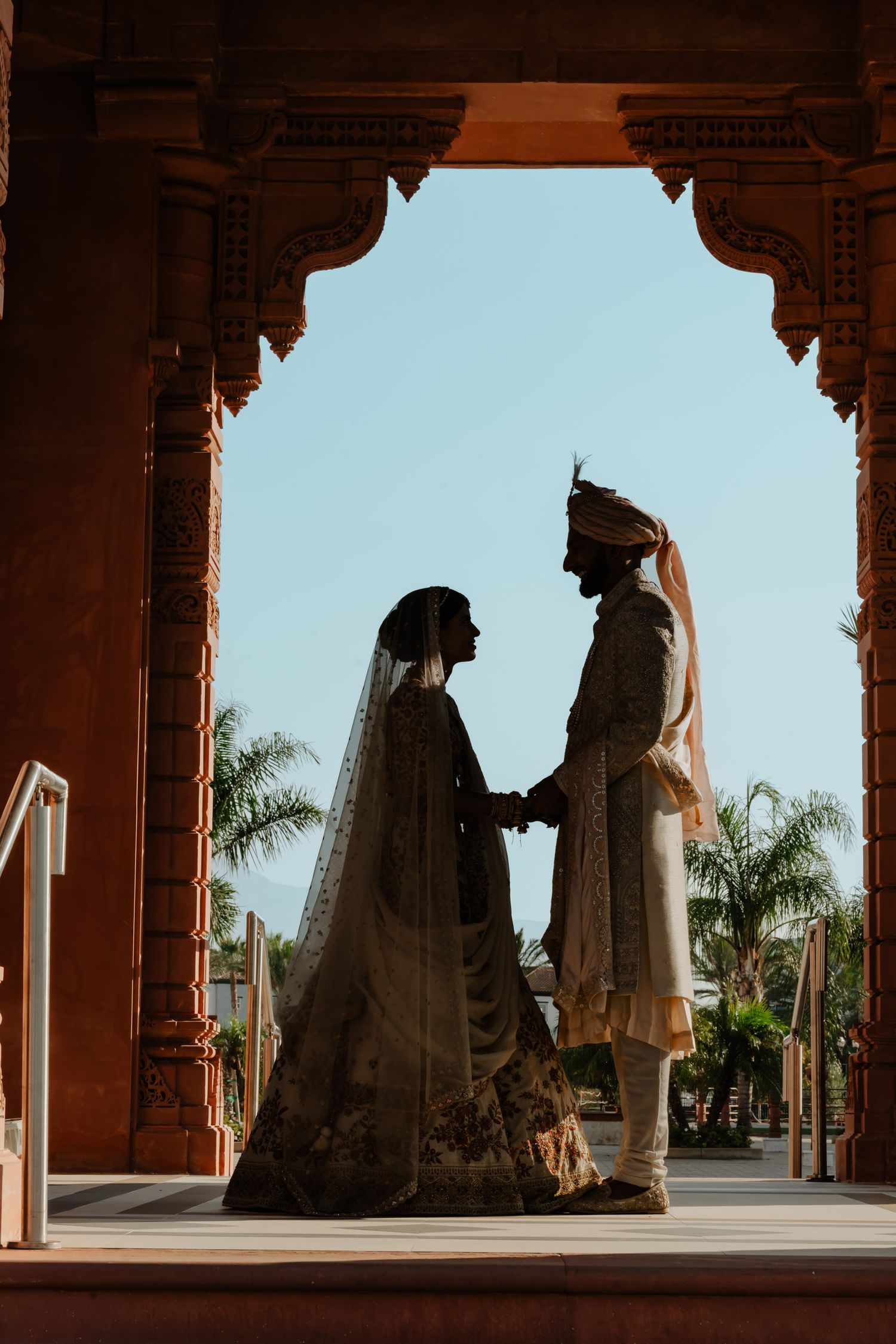 Silhouetted figures stand in archways against bright sky with palm trees.