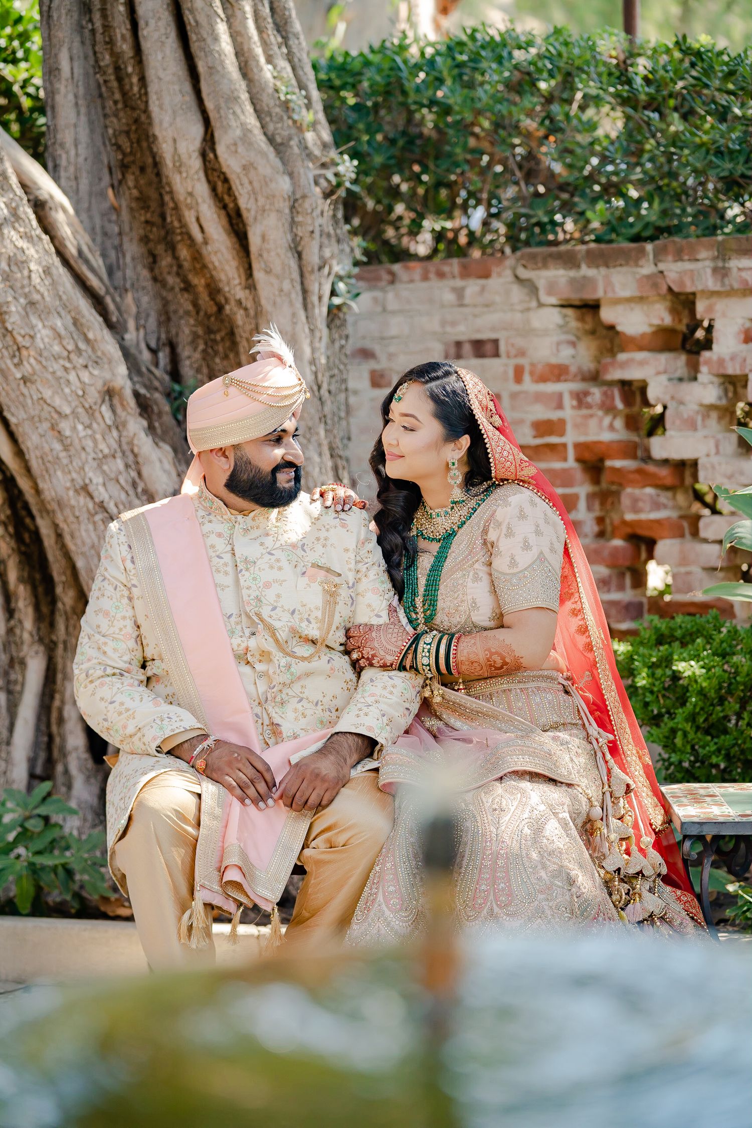 A couple in traditional Indian wedding attire sits together on a bench in a garden setting.