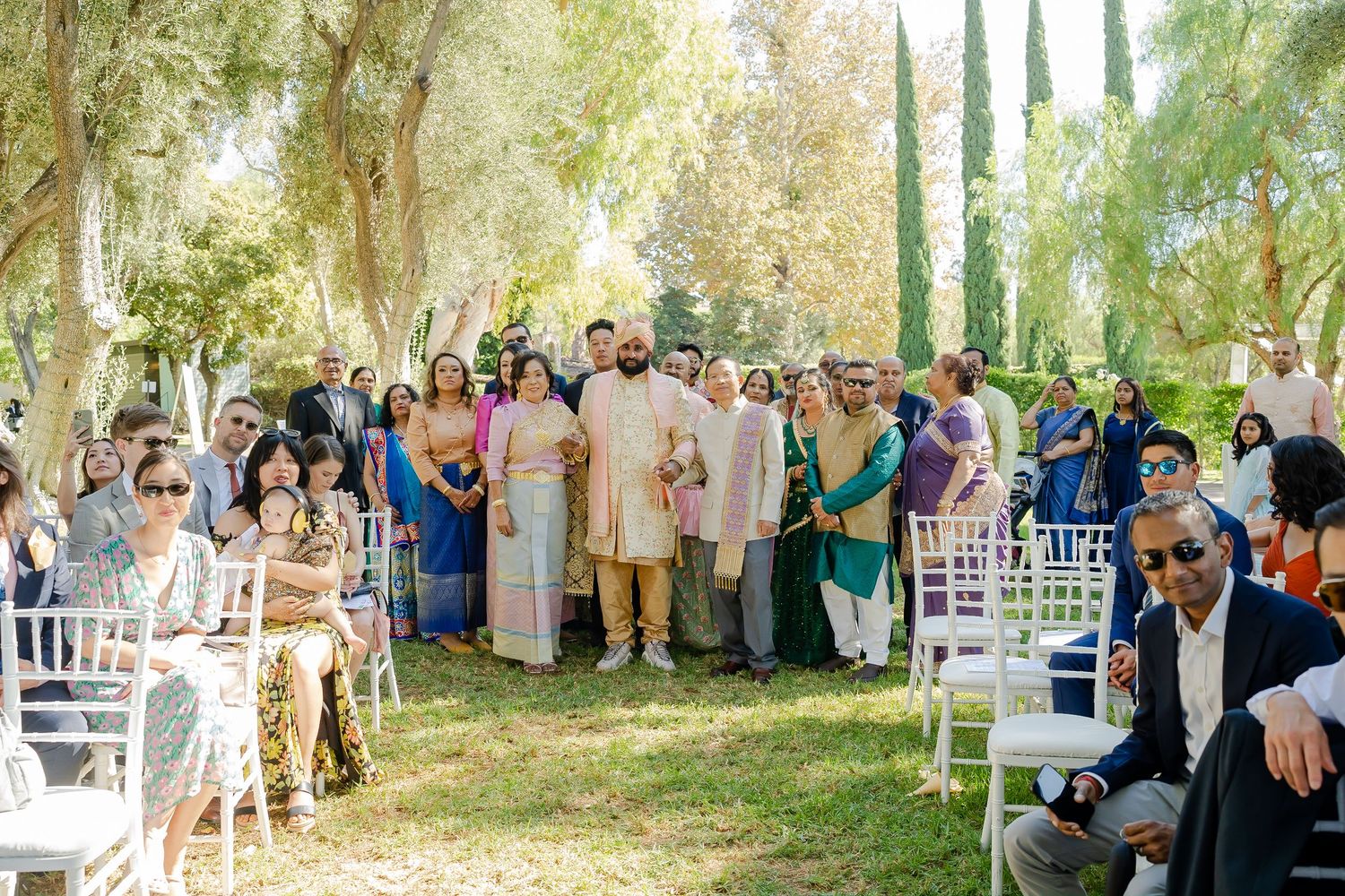 Guests gather under a canopy of trees for an elegant outdoor wedding ceremony with white chairs and natural decor.