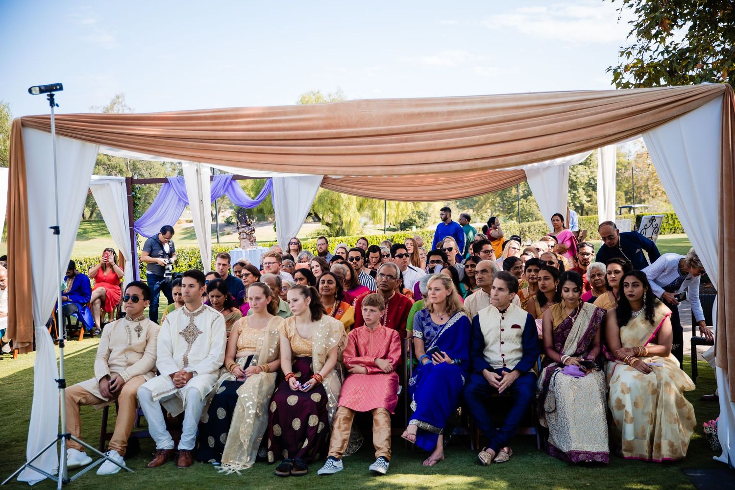 A panoramic view of wedding guests seated under a white tent canopy during an outdoor celebration ceremony.