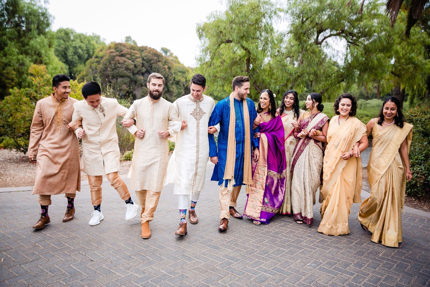 Wedding party members walk together outdoors wearing coordinated traditional South Asian attire in gold and neutral tones.