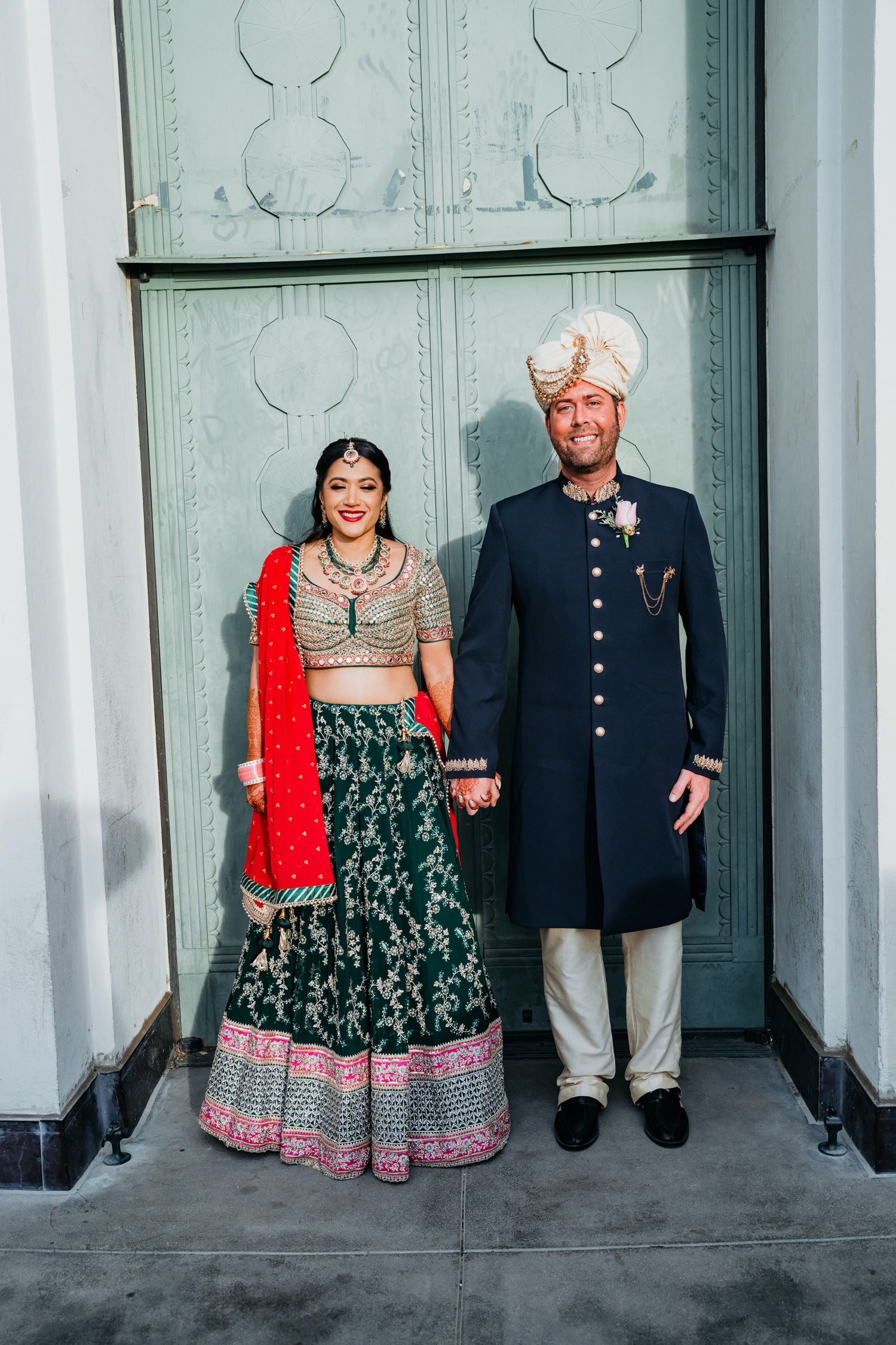Wedding couple in fusion attire posing against white columns and grey wall.