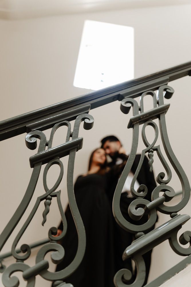 Couple in black outfits embracing on elegant staircase with ornate wrought iron railing.