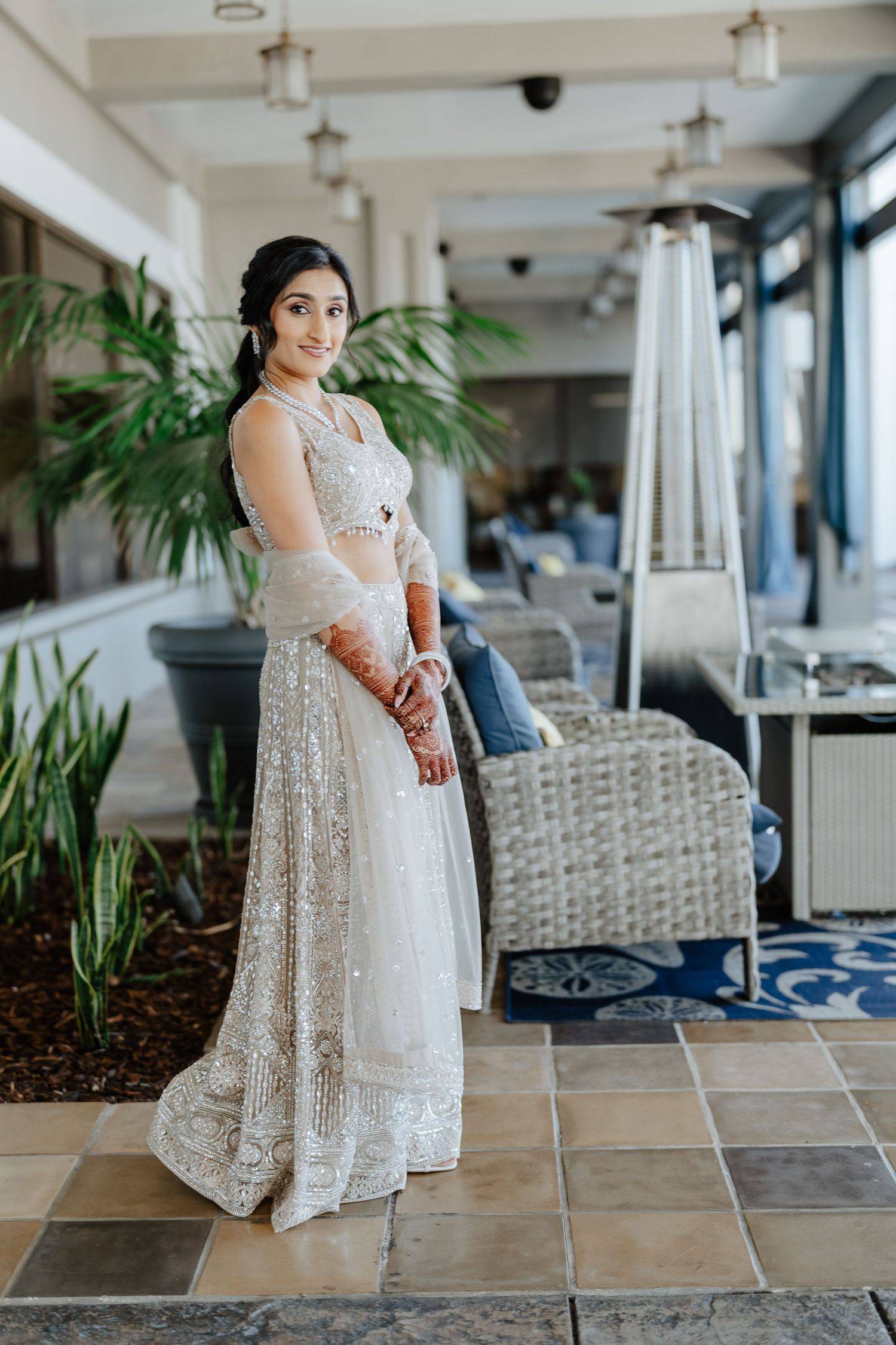 A bride in a white sequined lehenga with mehndi hands poses elegantly in a sunlit venue with tropical plants.