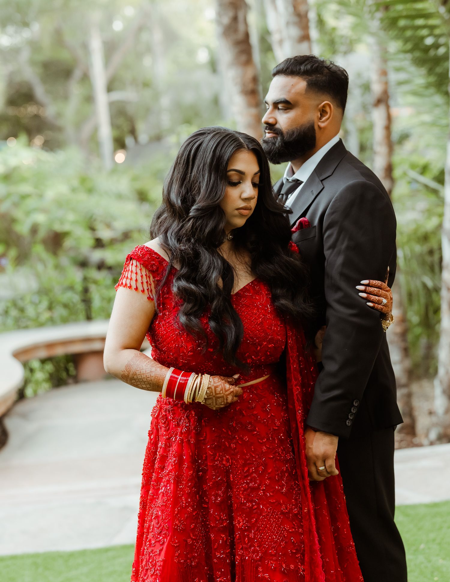 Couple in formal attire with red dress and dark suit posing in outdoor garden setting.