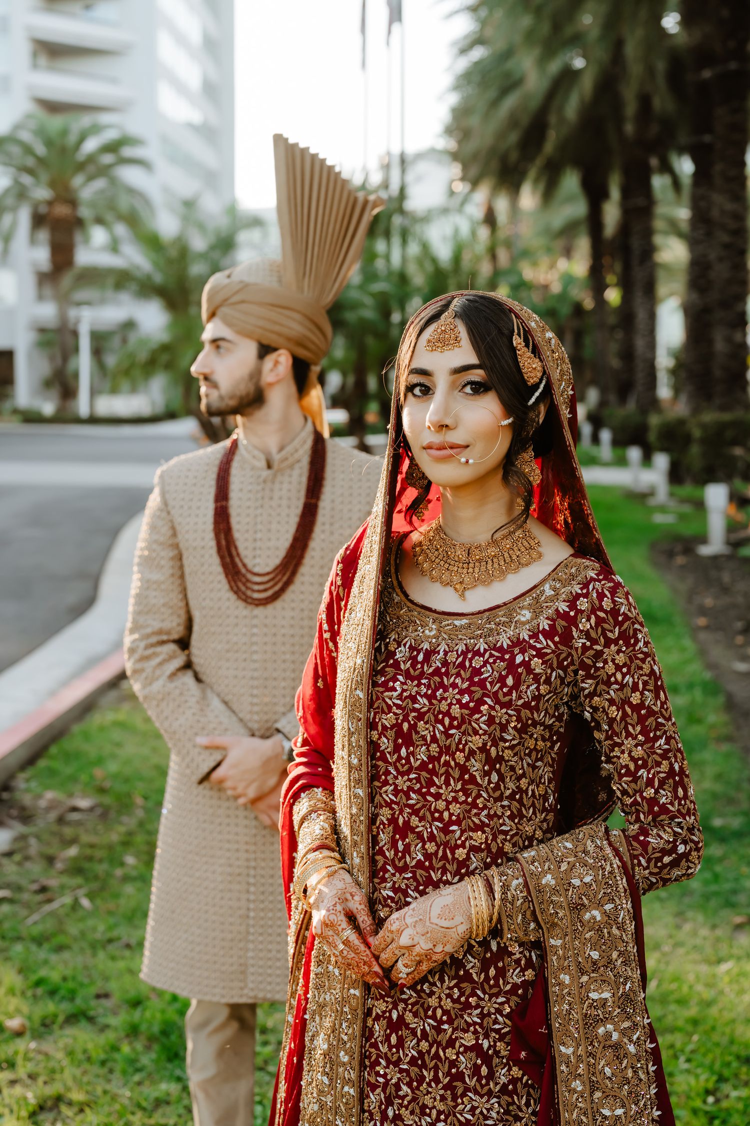 Traditional South Asian wedding attire featuring deep red embroidered clothing and gold accessories against a palm tree backdrop.