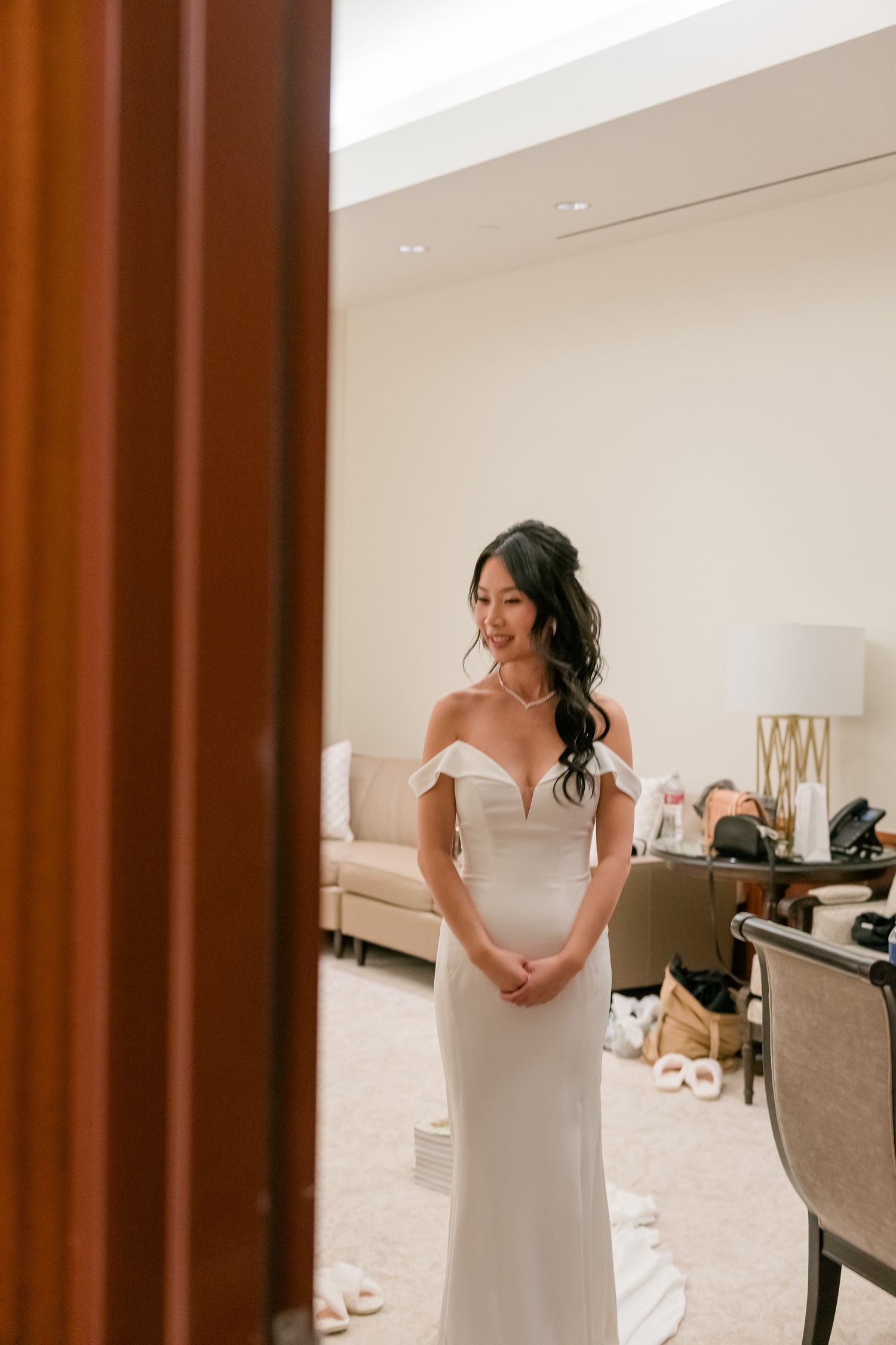 A bride in a white wedding dress gets ready in an elegant room with upholstered chairs.