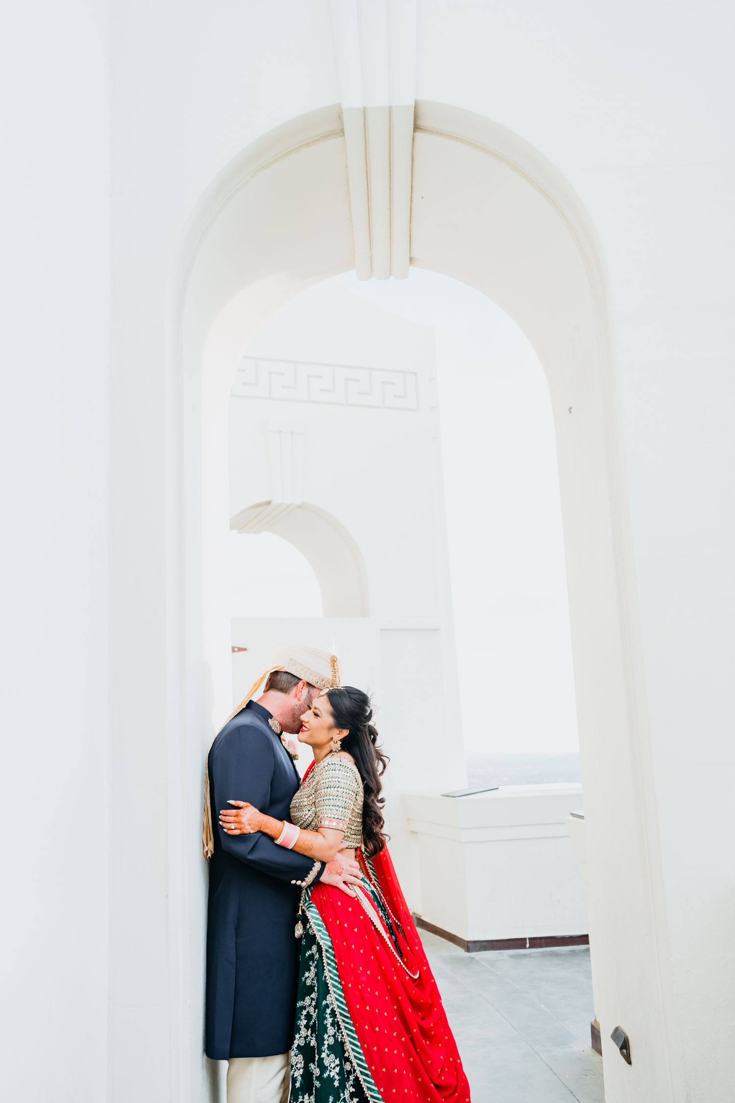 A couple embraces under a white archway with the bride wearing a red and gold traditional lehenga.