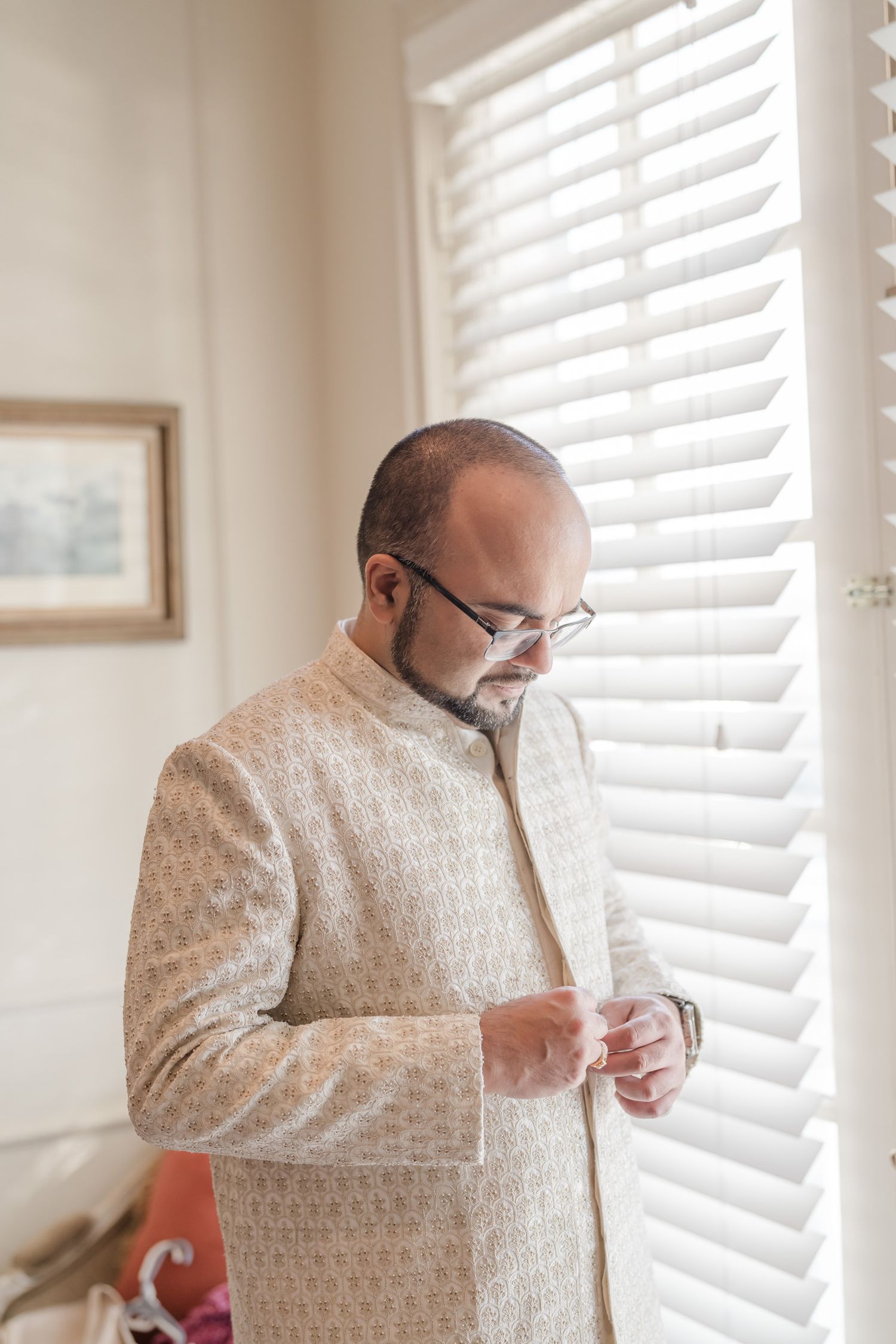 A person wearing a light colored traditional outfit adjusts their attire near a window with white blinds.