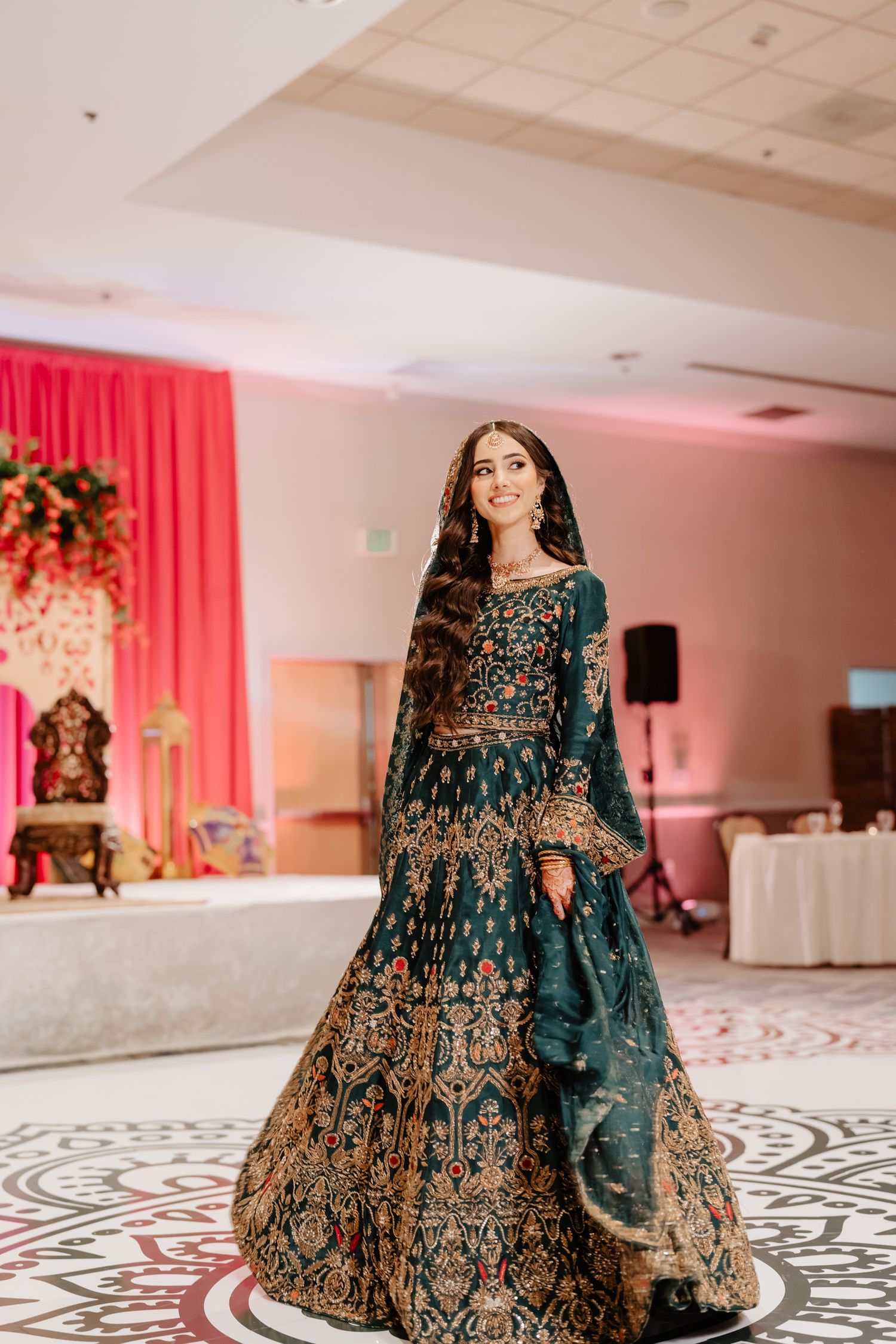 Ornate dark teal and gold embroidered lehenga dress on display in an elegant ballroom with pink decor.
