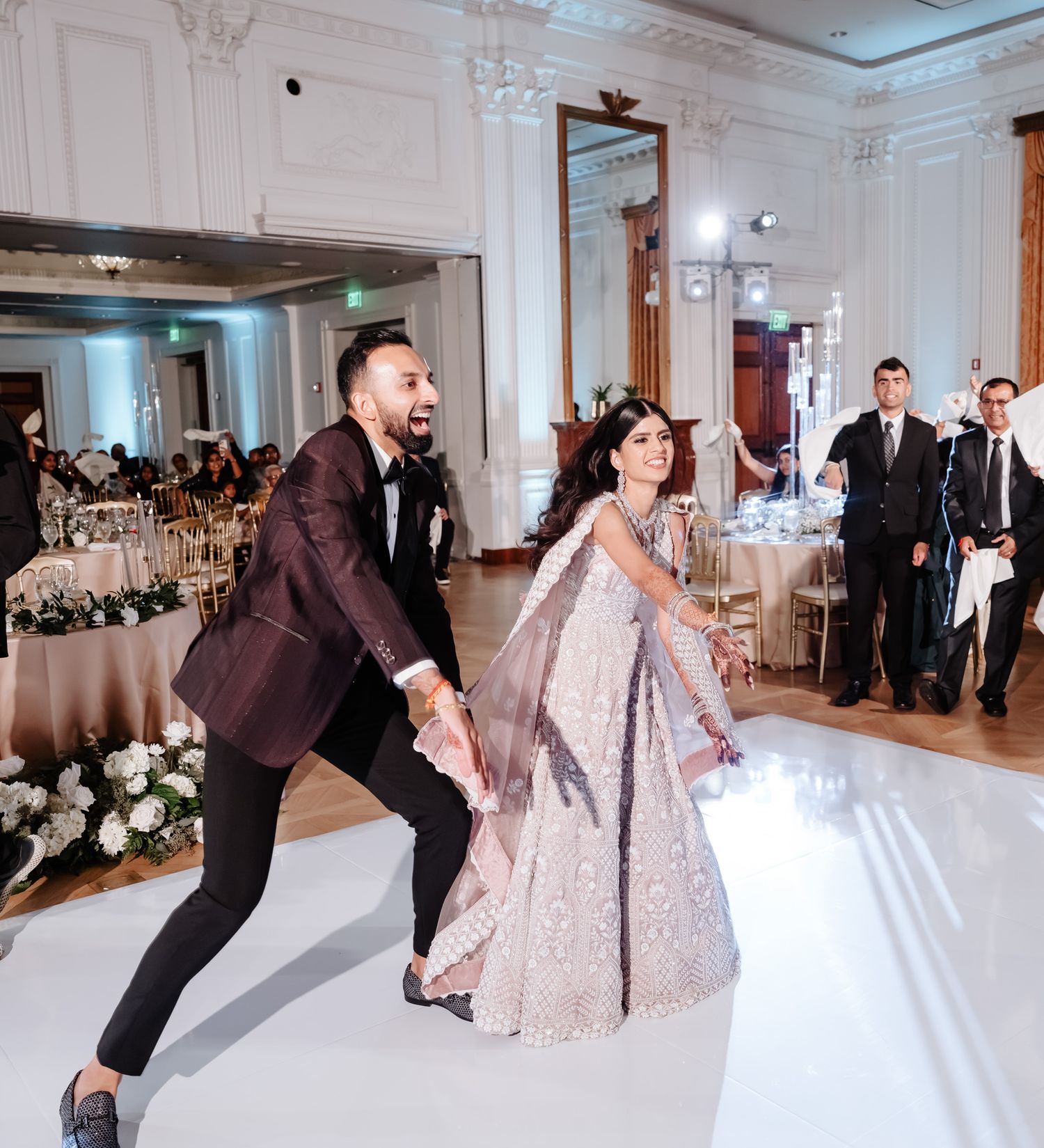 A couple dances together at an elegant wedding reception in a grand ballroom with ornate white decor and floral arrangements.