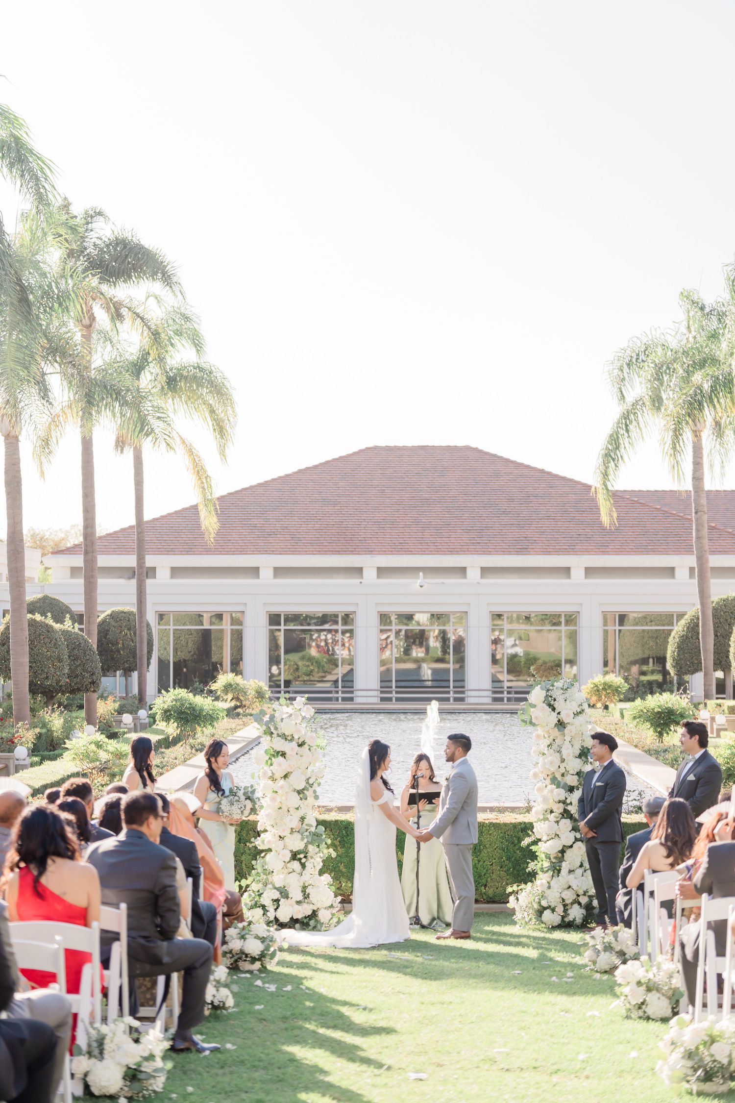 Outdoor wedding ceremony setup with white chairs and floral arrangements under palm trees.