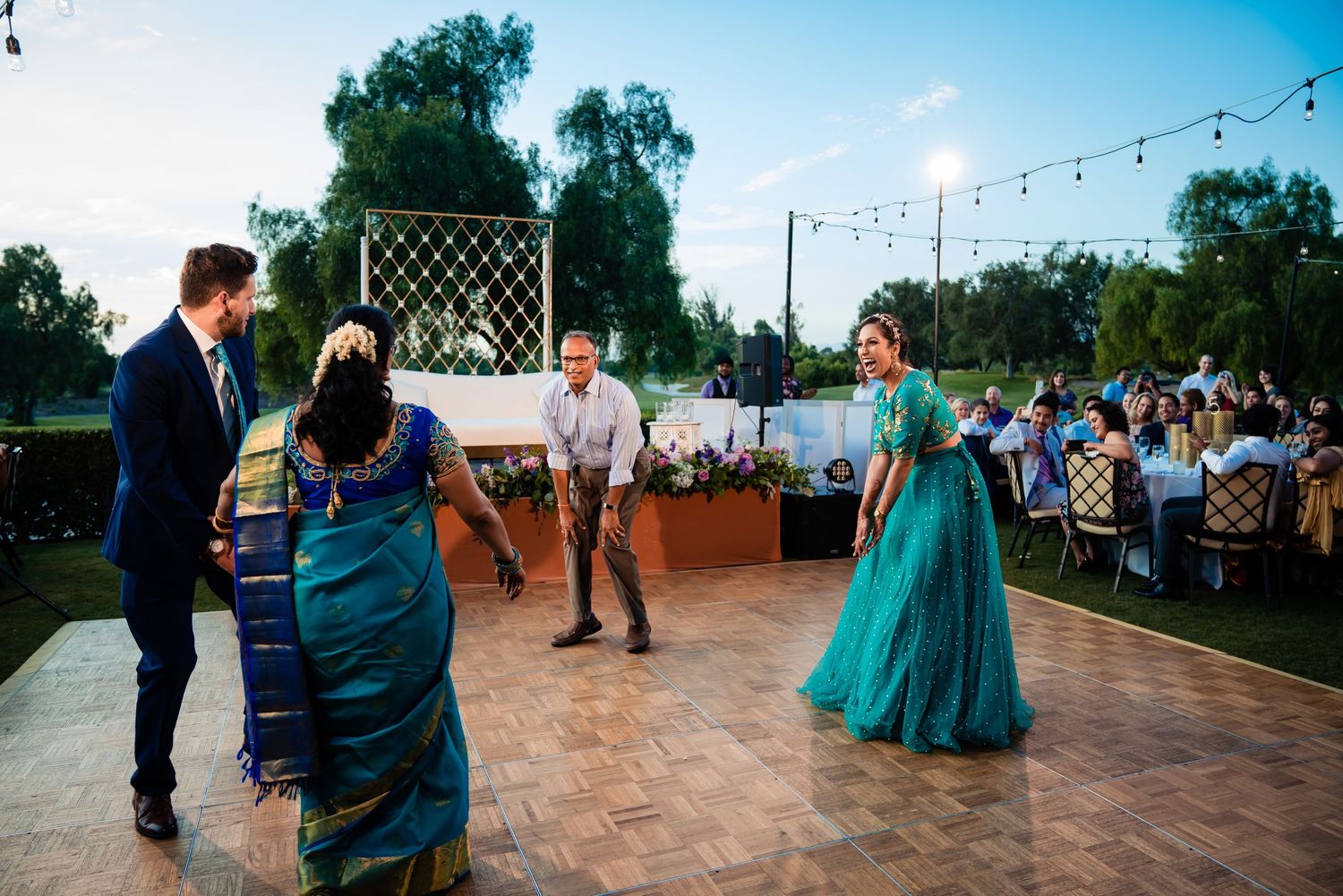 Outdoor wedding reception with guests dancing on a patio under blue skies and string lights.