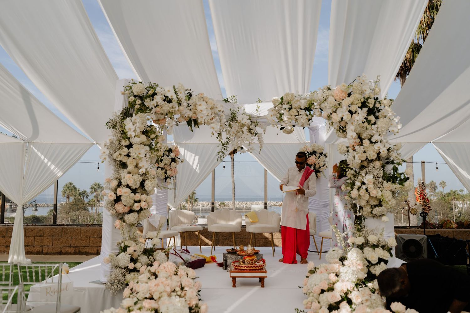 Elegant outdoor wedding mandap adorned with white and blush floral arches under a draped white canopy.