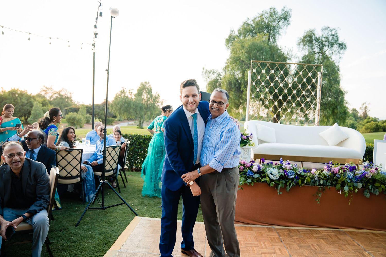 Guests dance and celebrate at an outdoor wedding reception under string lights and greenery.