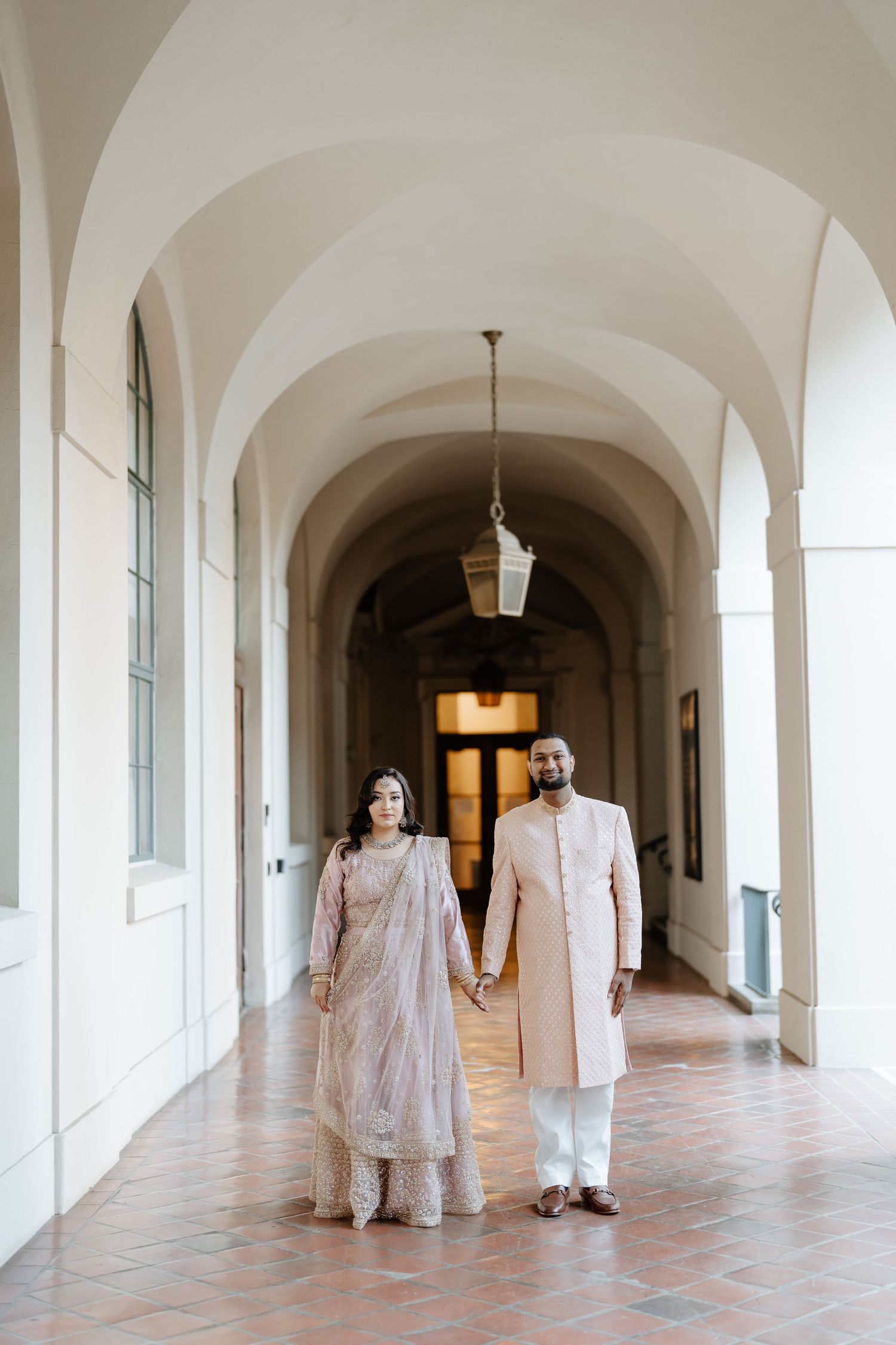 A couple walks through an elegant arched hallway wearing traditional South Asian attire.
