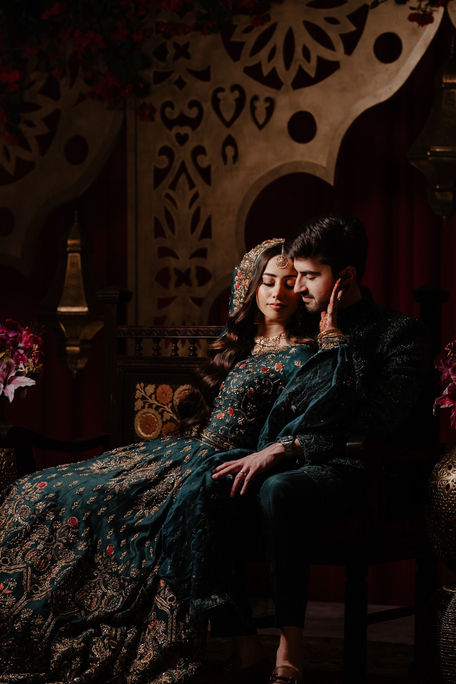 A couple in elegant dark attire reclines on a luxurious sofa in a dimly lit room with ornate wall decorations.