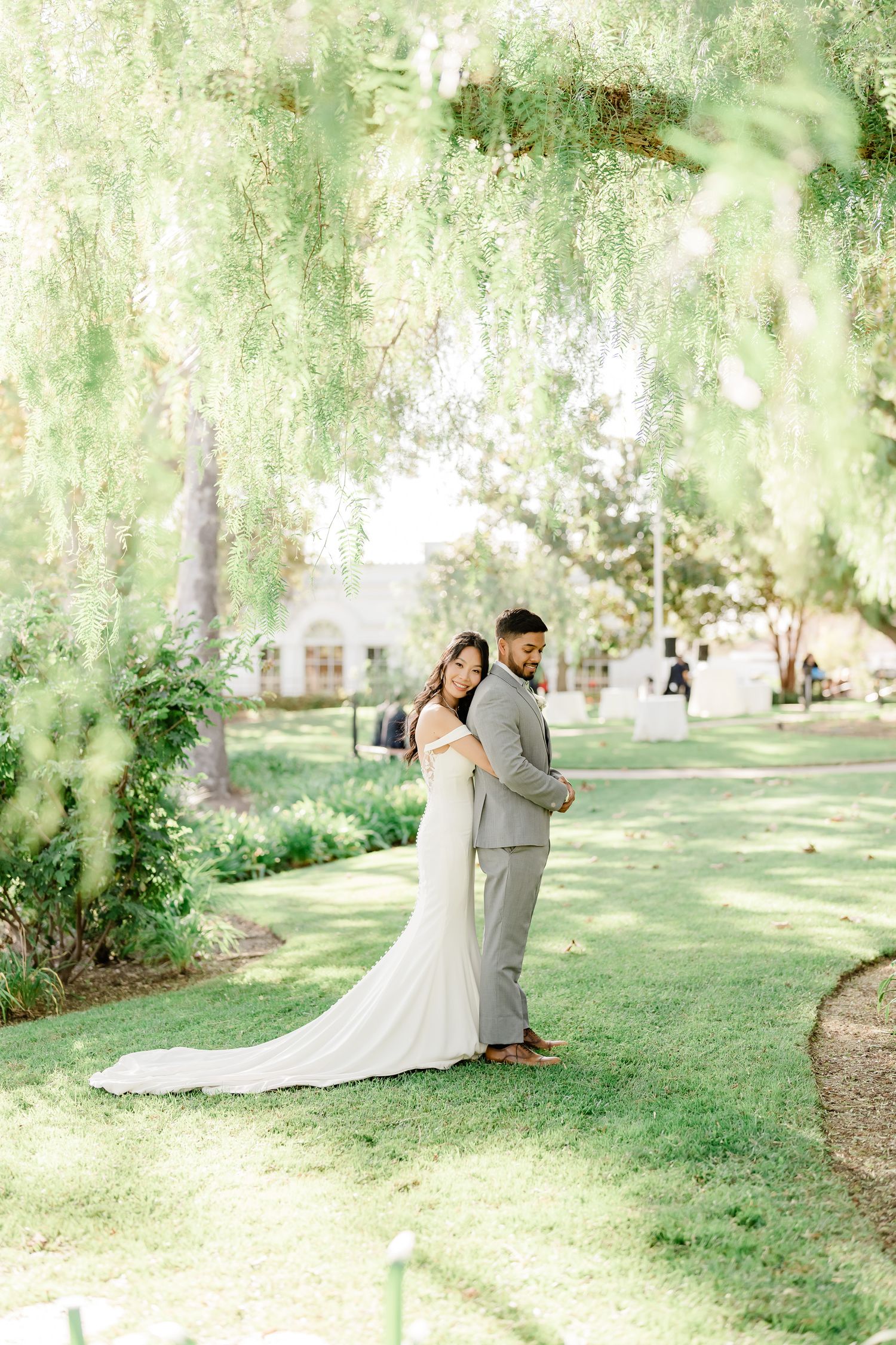 A couple shares an intimate moment under weeping willow trees in a sunlit garden setting.