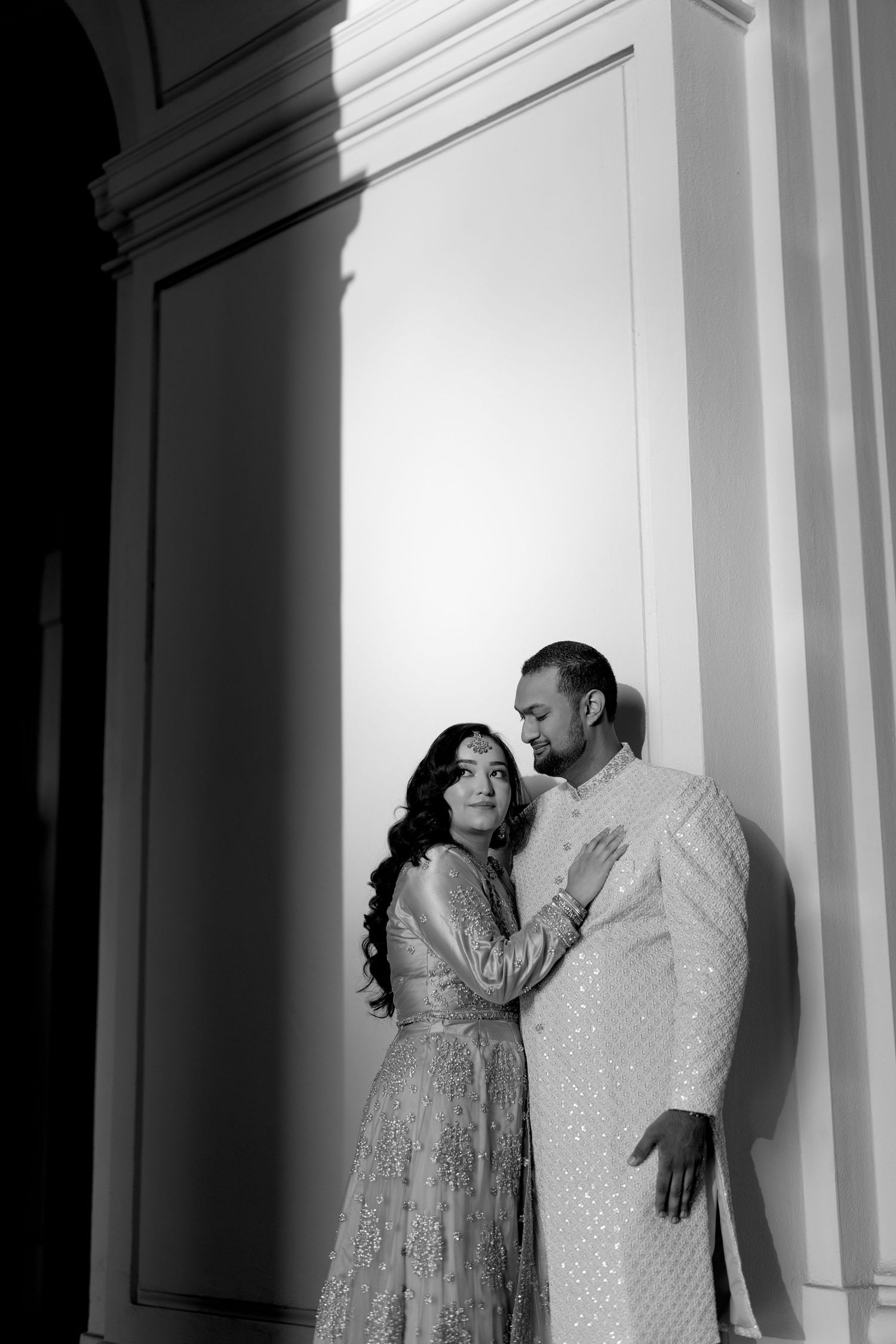 A couple in traditional South Asian attire share an intimate moment against a white architectural backdrop.