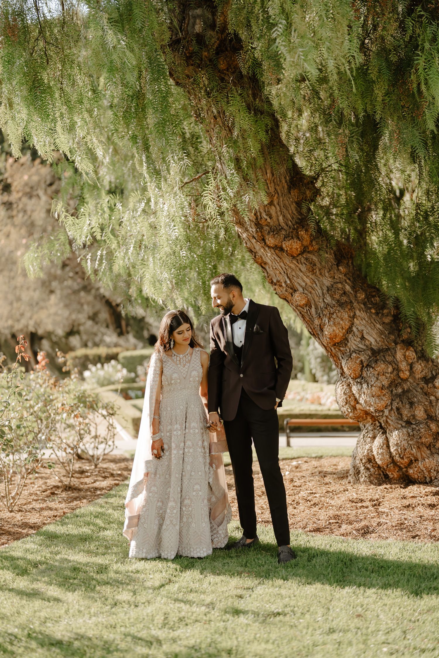 A couple in wedding attire poses under a dramatic weeping willow tree in an outdoor garden setting.