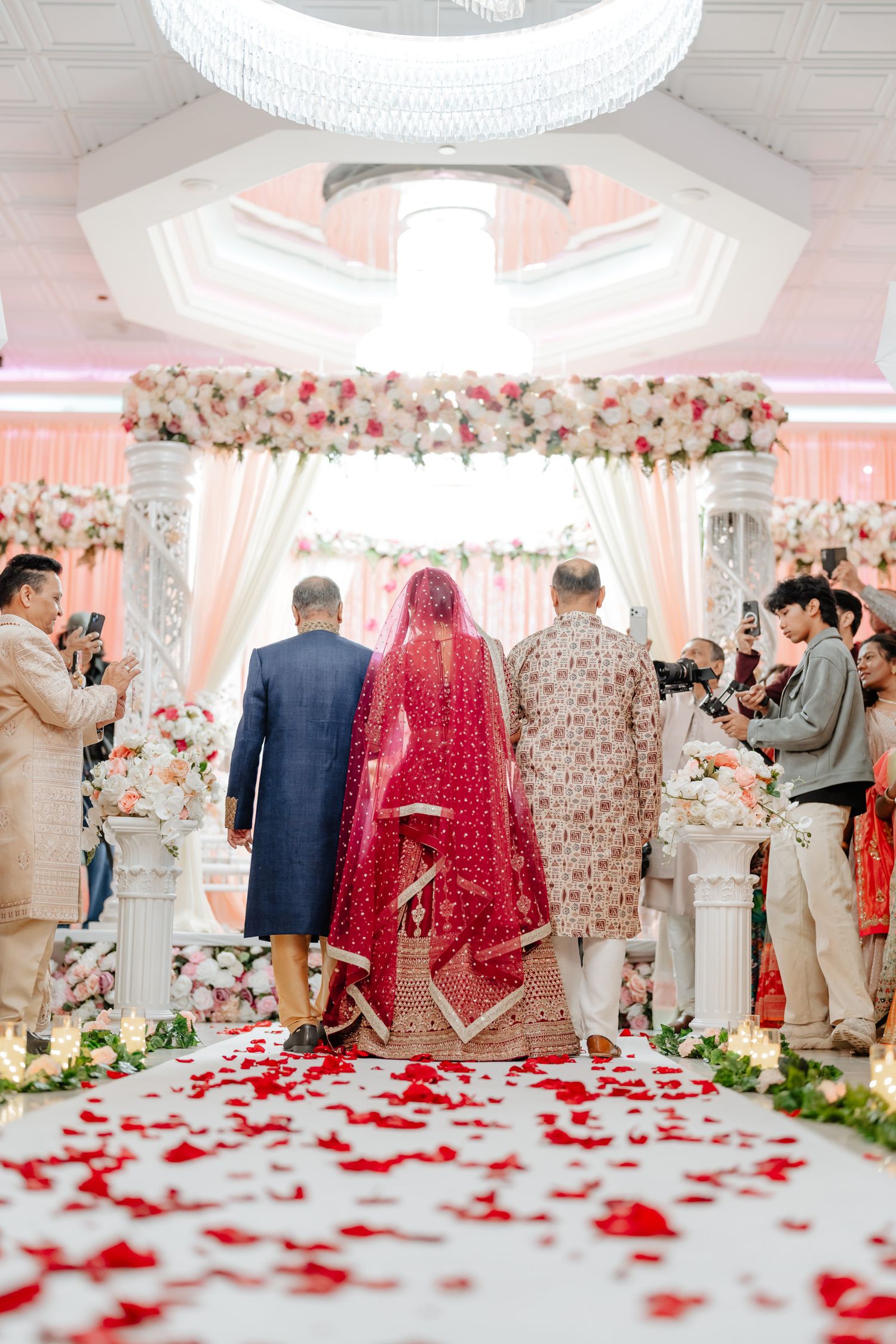 Wedding ceremony setup with floral arch and rose petals scattered on white aisle in elegant ballroom.