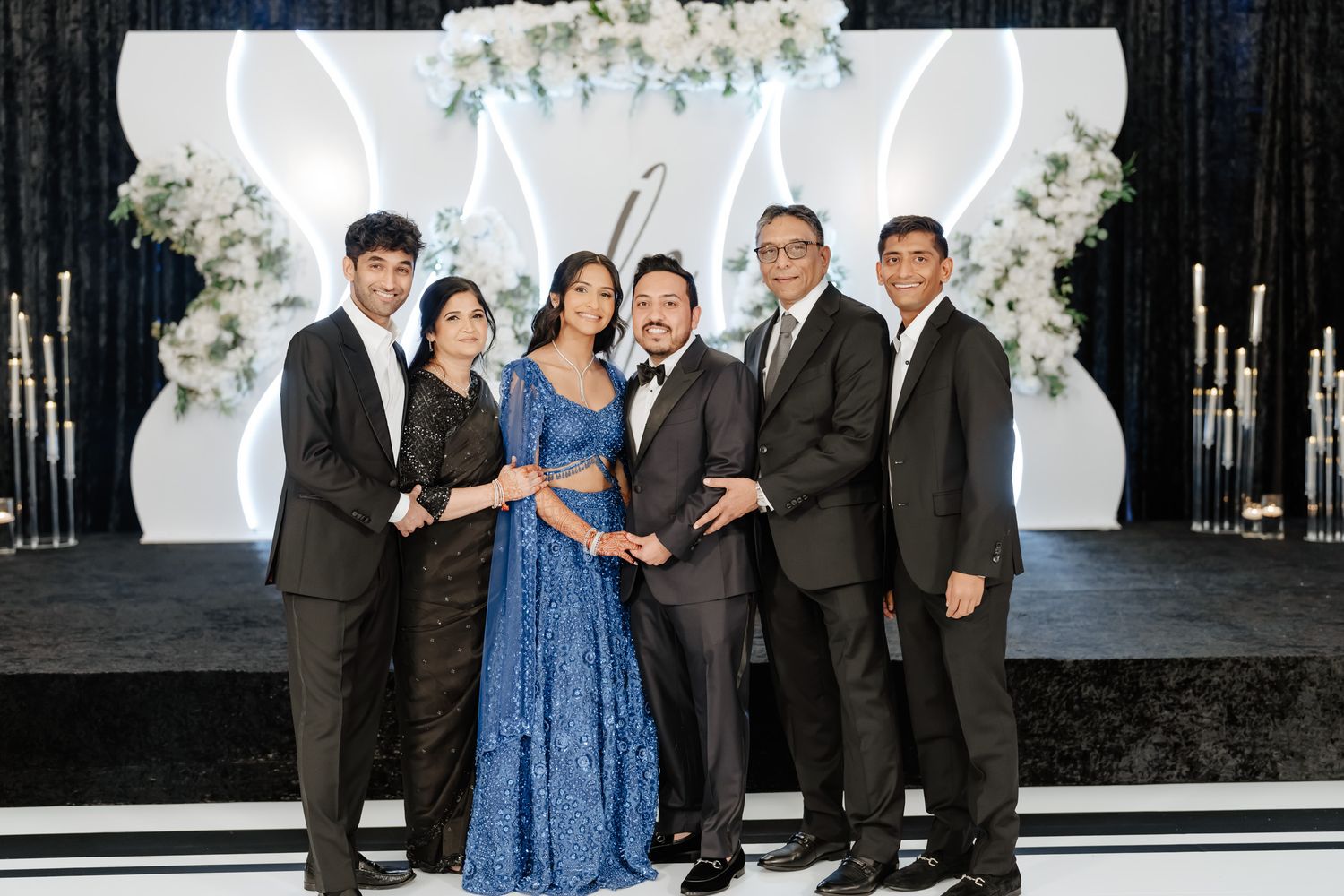 Wedding party group posing together in formal attire against white floral backdrop.