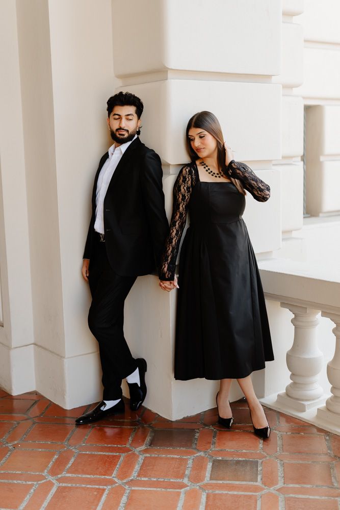 Stylish couple in matching black outfits holding hands while leaning against white wall on terrace.