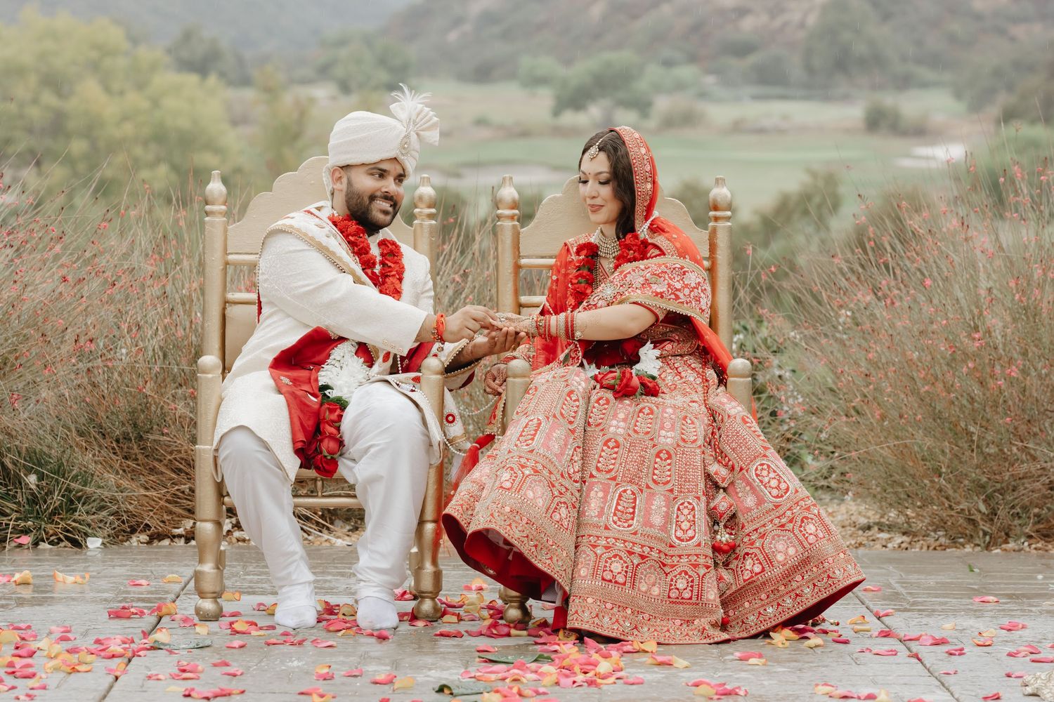 Indian bride and groom in traditional red and white wedding attire seated outdoors exchanging vows with rose petals.