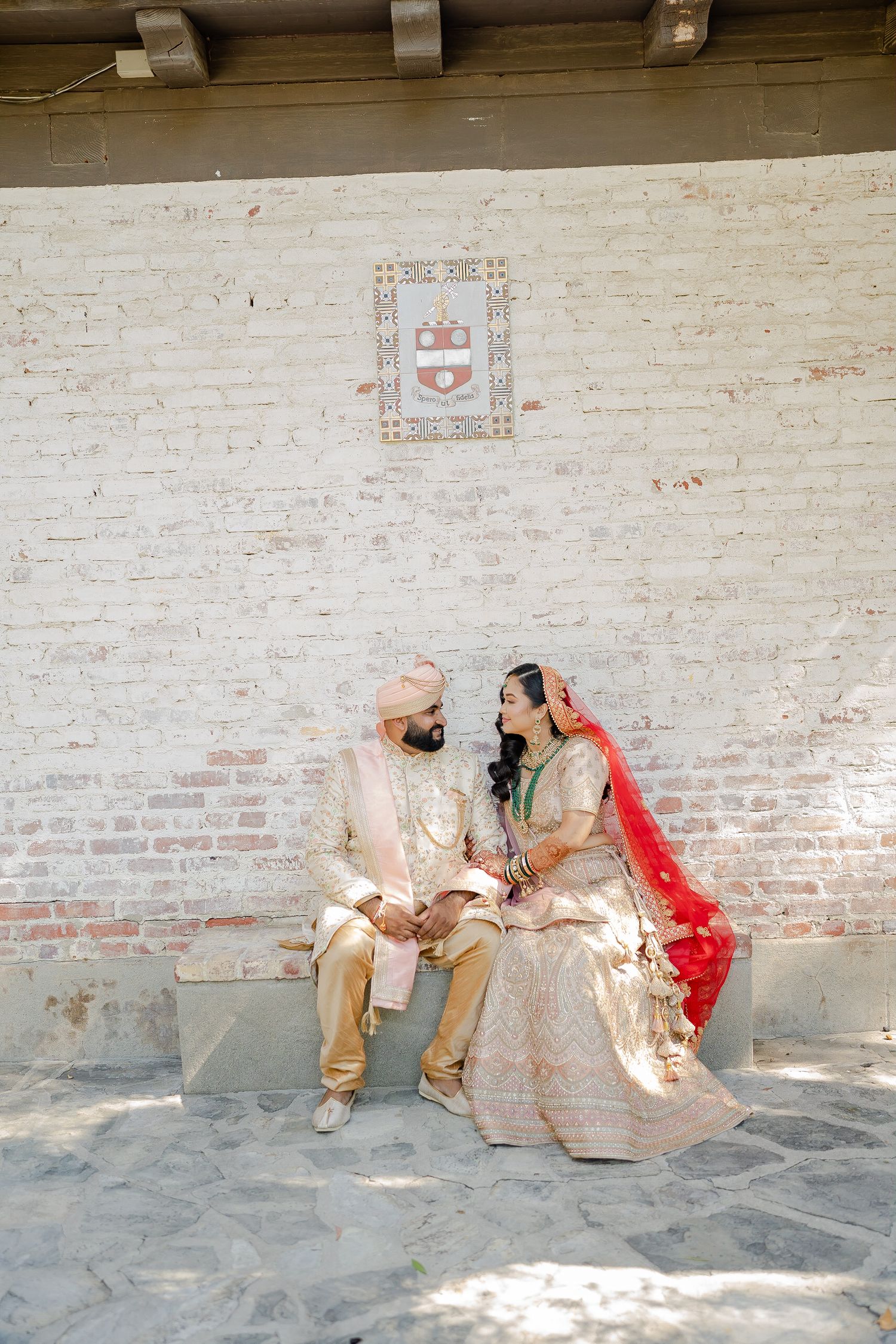 A couple in traditional South Asian wedding attire shares intimate moments against a rustic brick wall background.