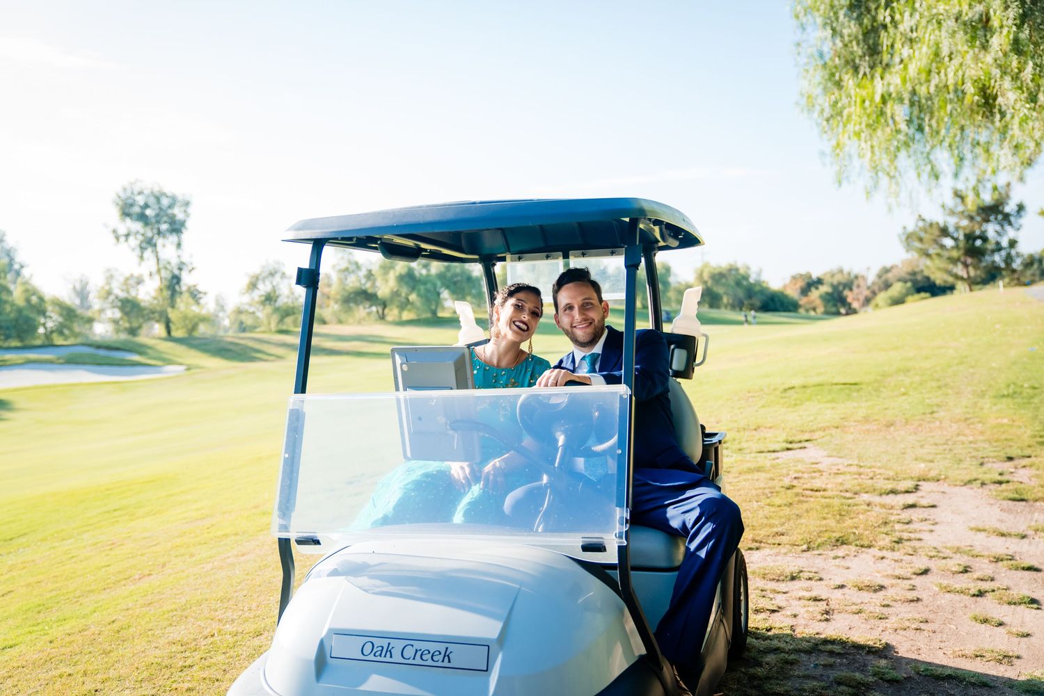 Wedding party riding in golf carts across a sunny golf course with trees in the background.
