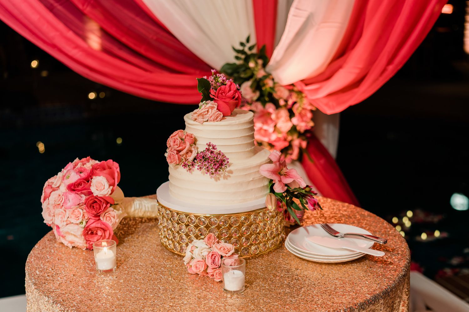 Wedding cake decorated with pink flowers and gold accents on a sequined tablecloth.