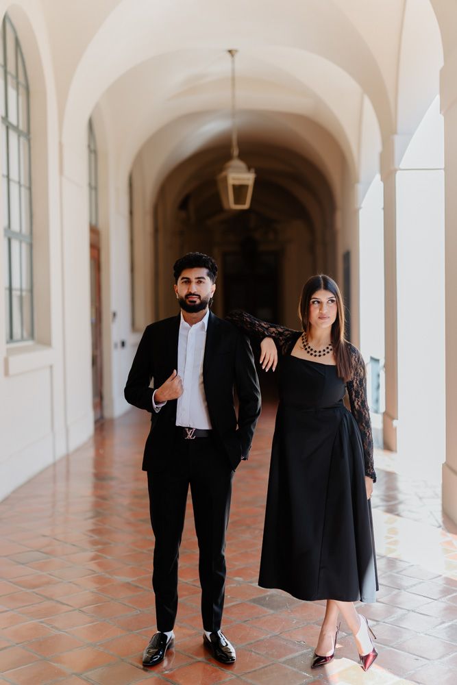 Stylish couple in black outfits walking through sunlit arched hallway.