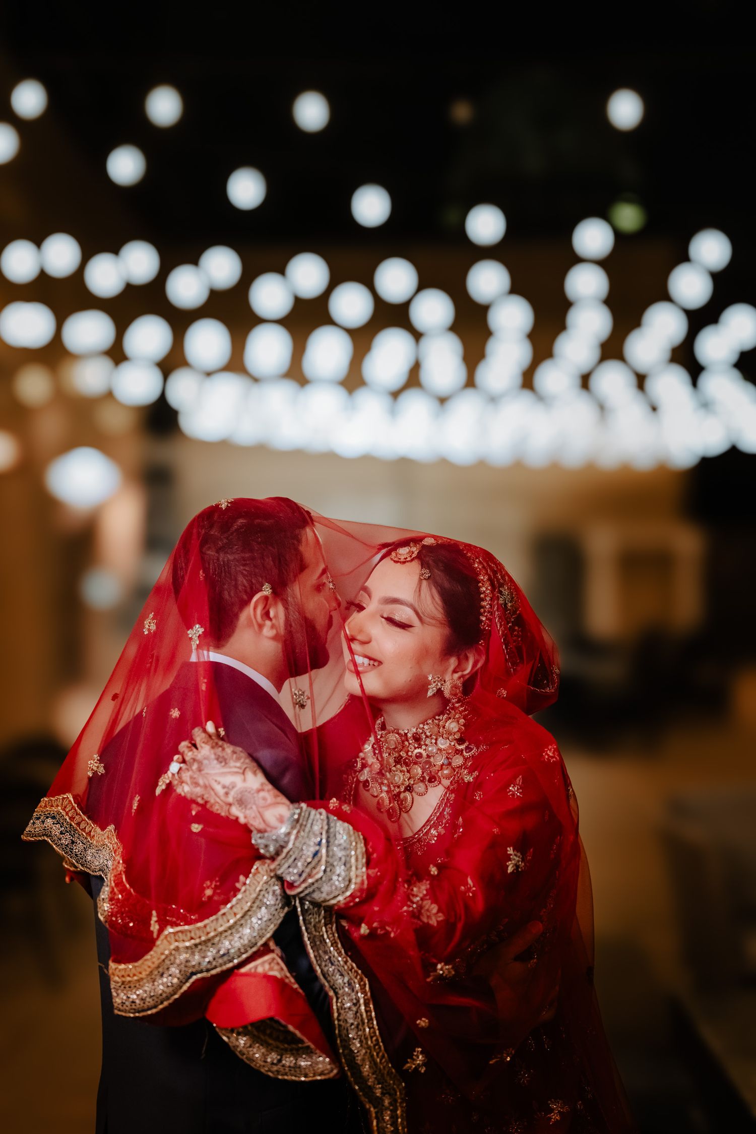 A couple shares an intimate moment under a flowing red veil with twinkling lights in the background.