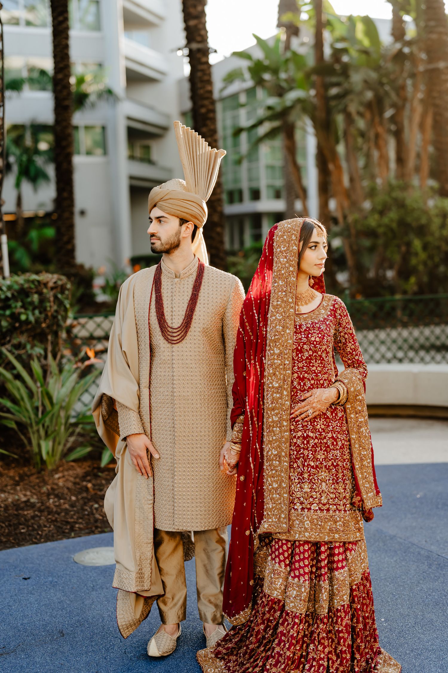 Traditional South Asian wedding attire featuring beige and red embroidered outfits against a palm tree backdrop.