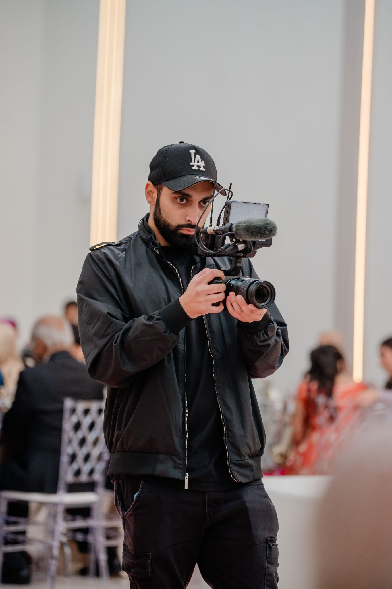 Videographer in LA cap holds camera gimbal stabilizer at indoor event.