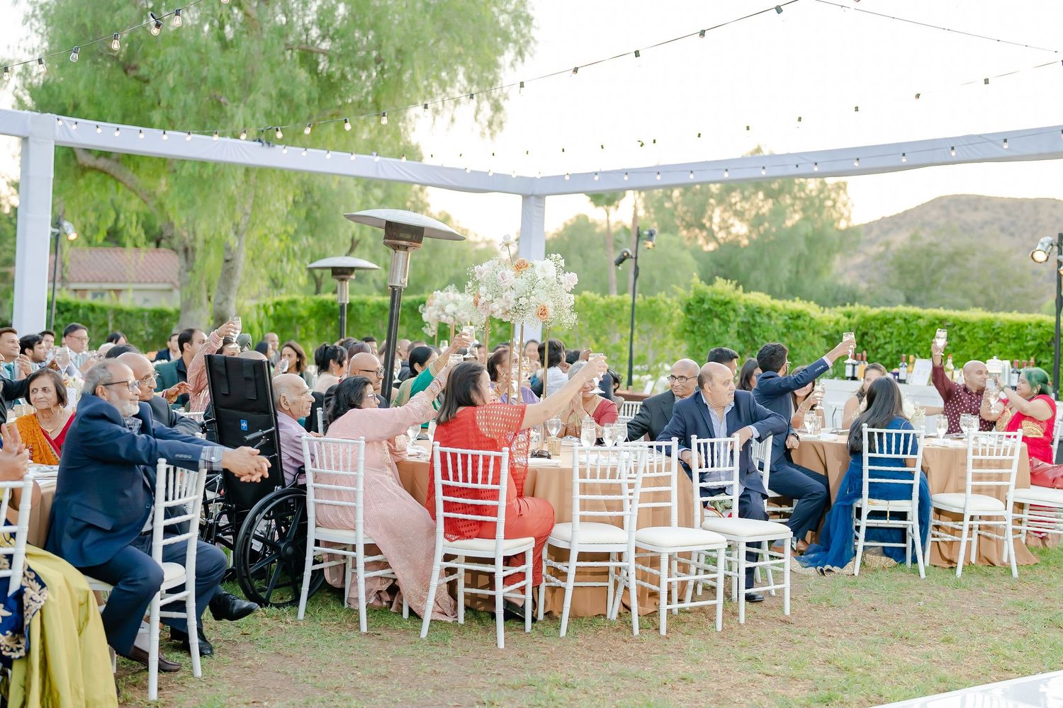 Wedding guests seated at round tables with white chairs under string lights and trees at an outdoor venue.