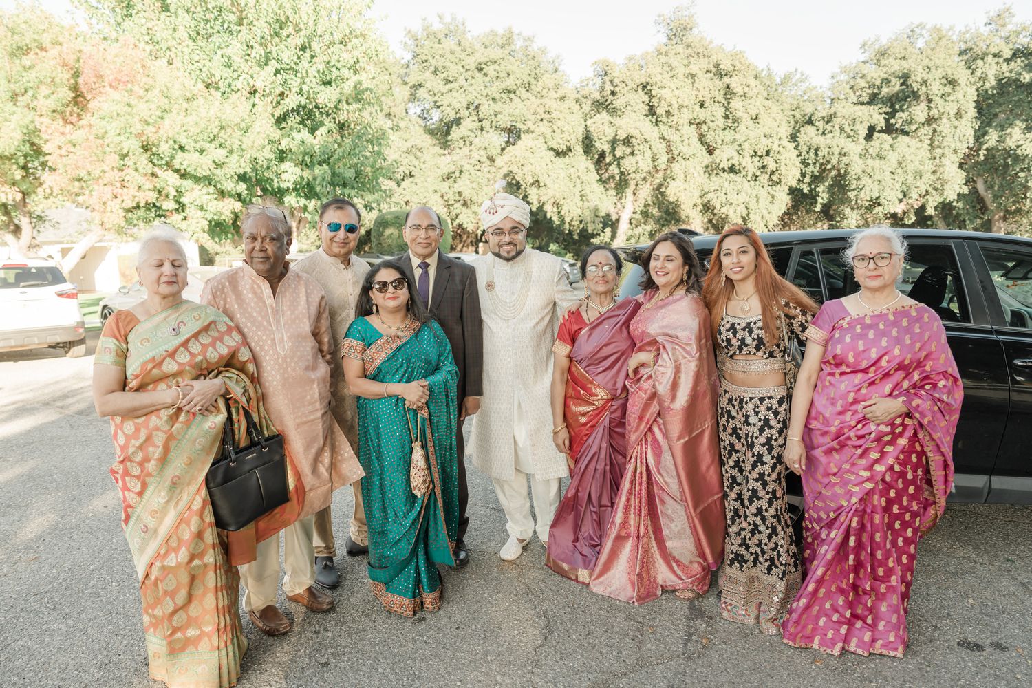 Wedding guests in vibrant saris and traditional Indian clothing pose together outdoors under trees.