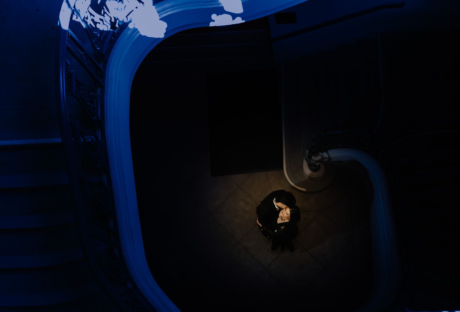 Aerial view of a lone figure illuminated by spotlight in a dark blue architectural space with curved walls.